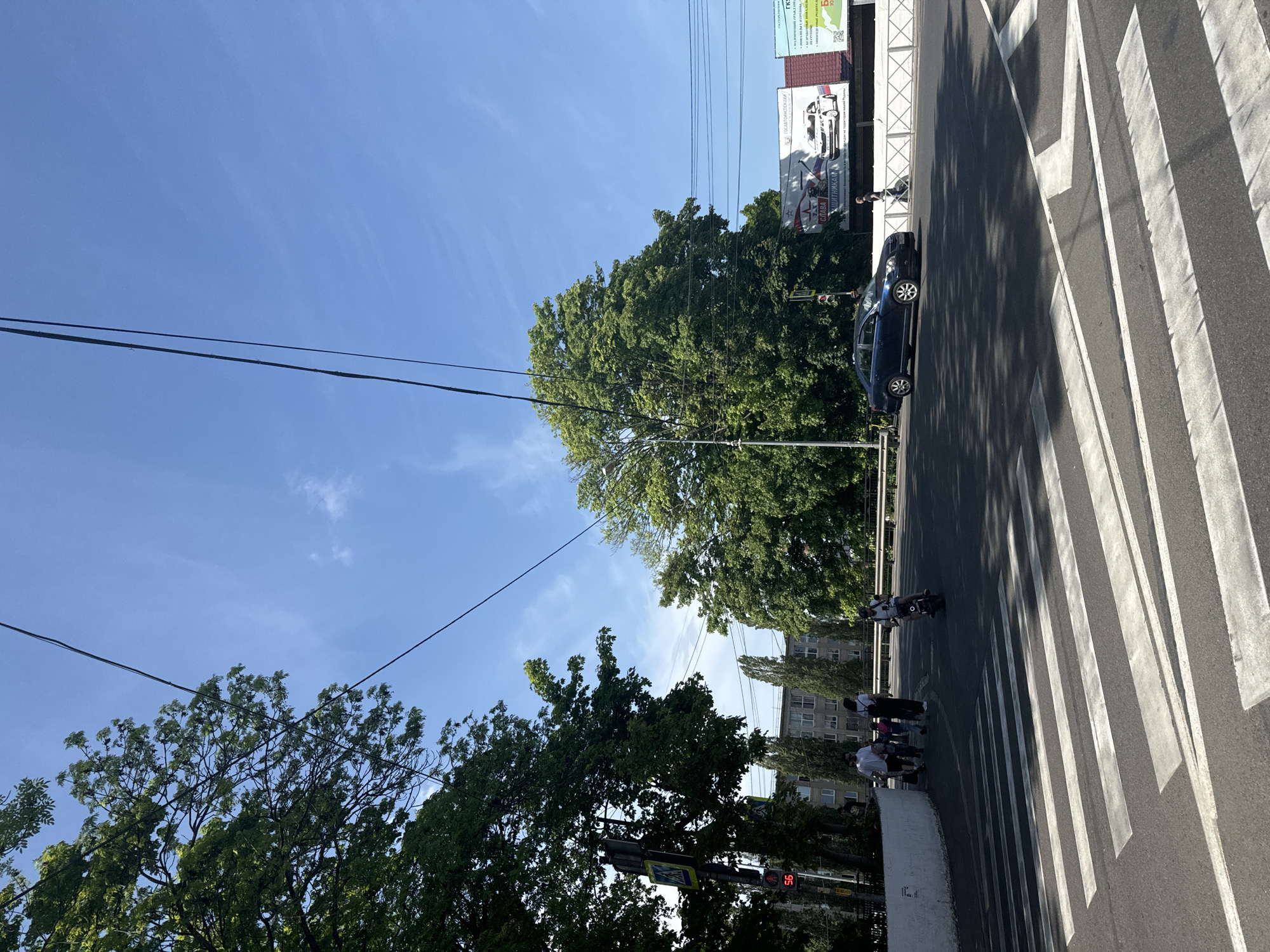 The image captures a street scene in an urban setting. A zebra crossing dominates the foreground, with stark white stripes contrasting against the dark asphalt of the road. Pedestrians can be seen crossing the street, including a group of people and a person on a motorbike. A dark blue car is parked along the roadside.
Overhead power lines stretch across the clear blue sky. Lush green trees line the street, partially obscuring the buildings behind. A billboard is visible above the trees, displaying text and images. A traffic light is present at the intersection, indicating a controlled crossing.