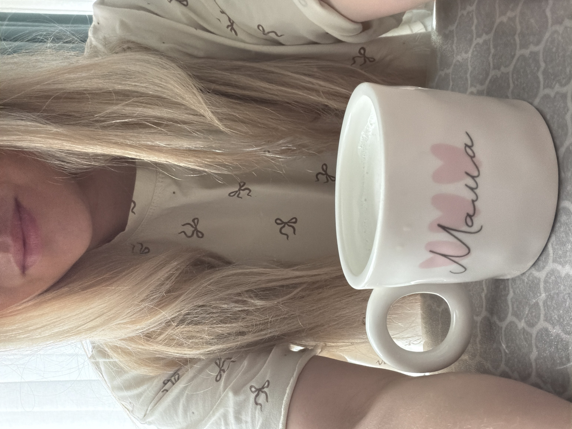 A selfie-style image shows a person with long, blonde hair lying down. The person's lips are visible in the left portion of the frame. They are wearing a light-colored shirt with a pattern of small, stylized bows. To the right of the person is a white mug with the word "Mama" and pink heart shapes printed on it. The mug is on a gray tablecloth with a geometric pattern. The location is not discernible from the image.