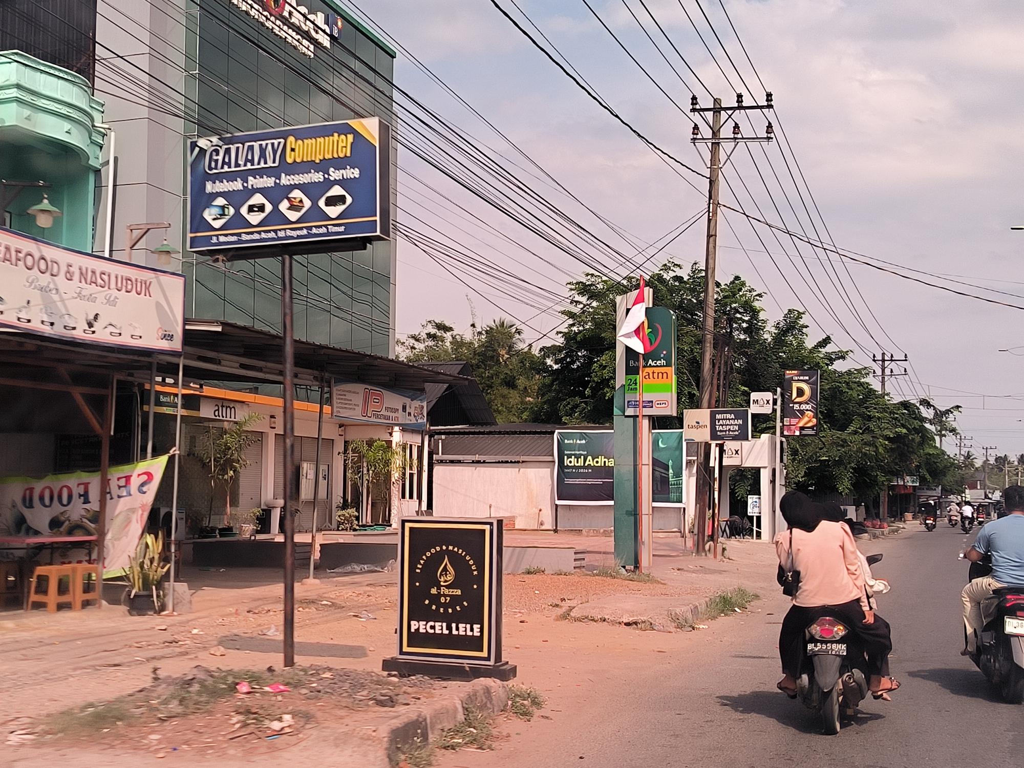 The image depicts a street scene in Seuneubokbacee, Indonesia. On the left, a sign advertises "GALAXY Computer," along with "Seafood & Nasi Uduk." An ATM kiosk is visible next to a "Fotocopy Percetakan & ATK" store. The middle ground shows signage for "Bank Aceh" and "Idul Adha." A "Pecel Lele" food stall sign is on the road curb. Two people riding a motorcycle are in the foreground. Another motorcycle and riders are further ahead, along with utility poles and wires overhead. A "Taspen" sign is also visible.