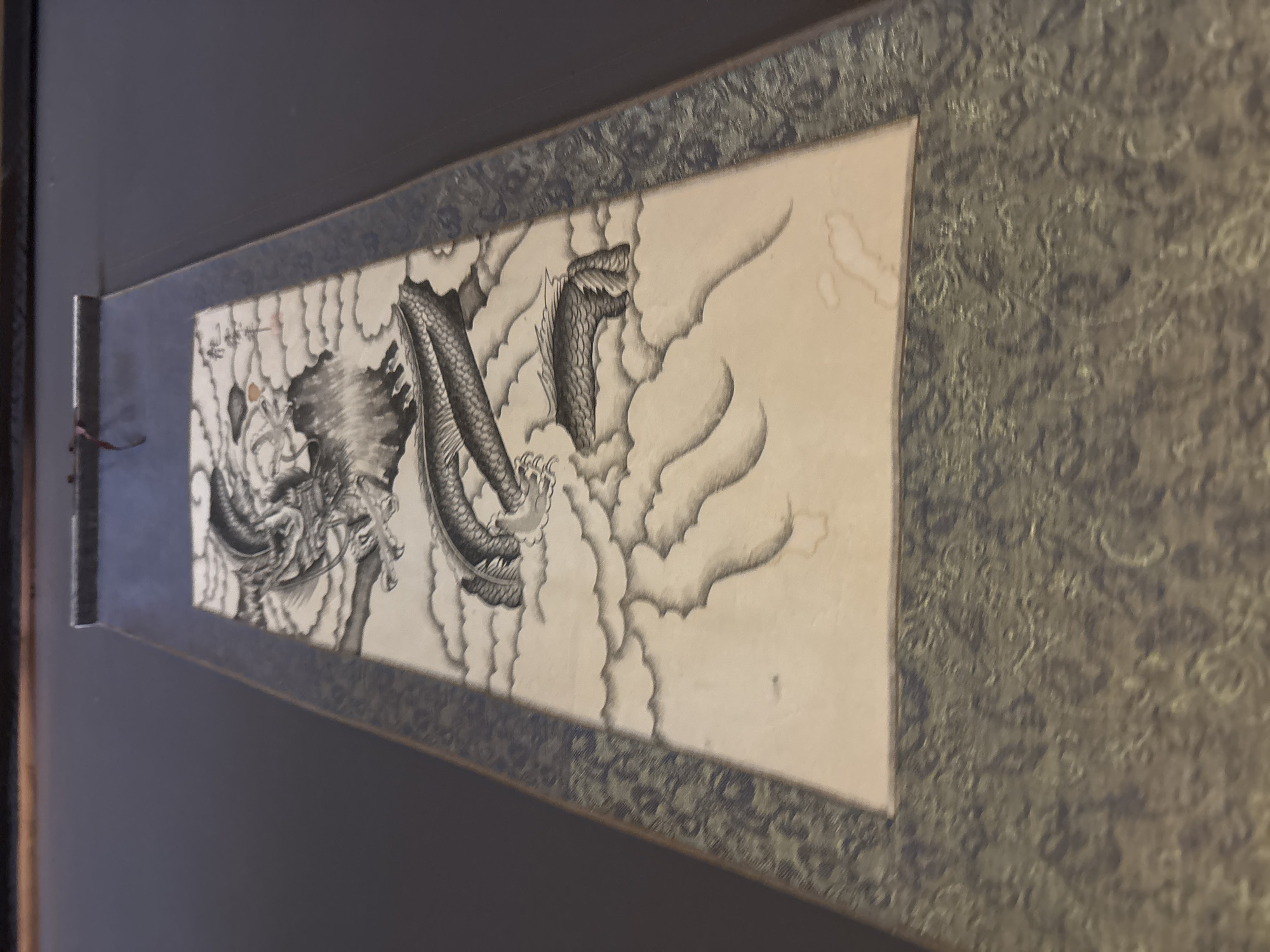 This is a detailed, forensic, matter-of-fact description of a scroll painting featuring a dragon. The painting depicts a dynamic, black and white dragon with a long, scaled body, sharp claws, and a fierce expression, rendered in ink on what appears to be aged paper. The dragon is depicted amidst swirling clouds, painted with varying shades of gray to create depth and movement. There are some light brown water stains visible on the paper, suggesting age or exposure. The scroll is framed by a dark, textured border, possibly silk or fabric, with a subtle pattern. The scroll is unrolled and displayed vertically, hanging against a dark, uniform background. There is no visible signage or context that would place this object in Novaya Usman, Russia. The image focuses solely on the artwork and its presentation as a scroll.