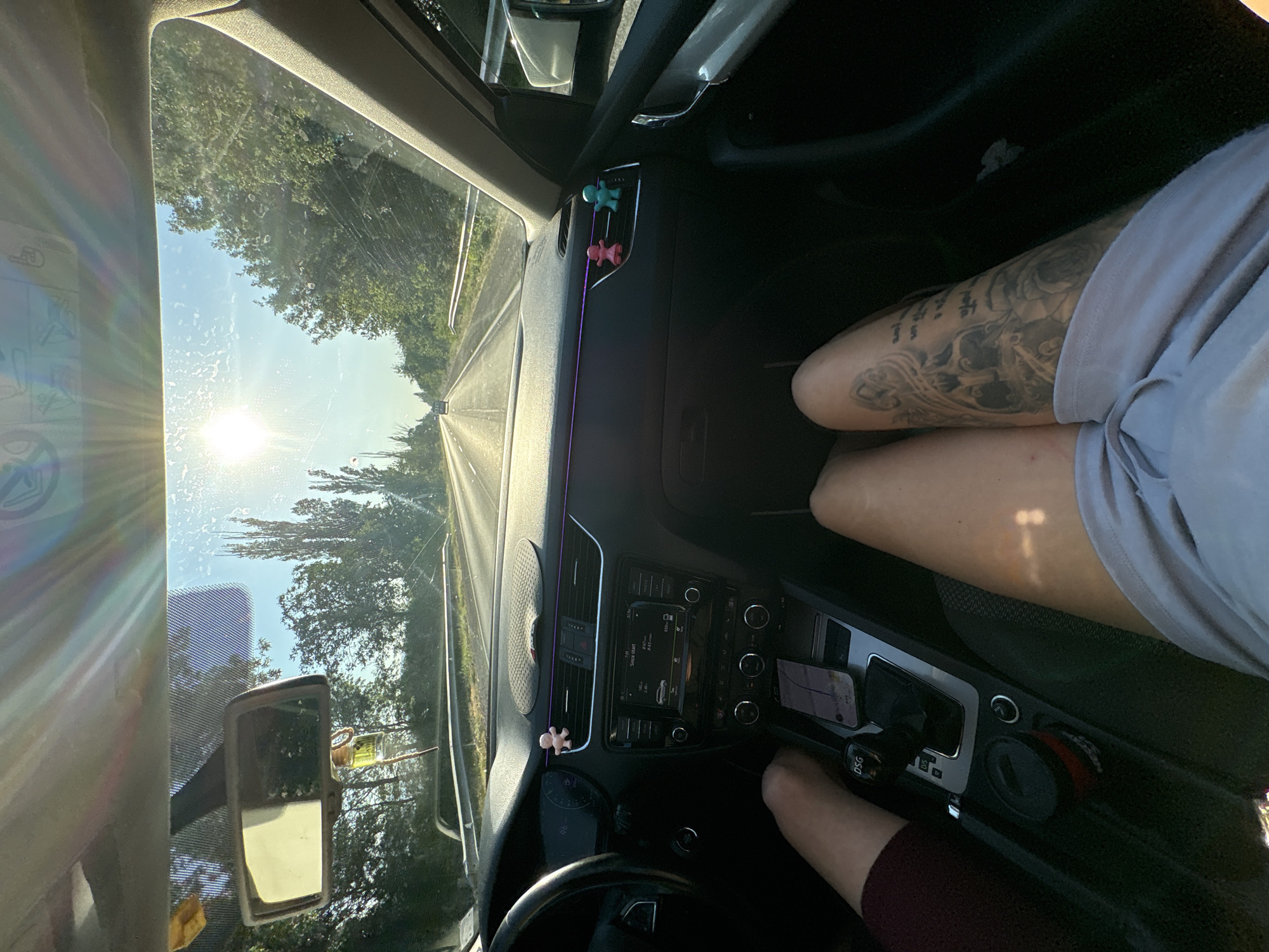 The image depicts the interior of a car traveling on a road, likely outside the city of Chintulovo, Bulgaria, judging by the sunlight and lush greenery. Two individuals are present; one's legs are visible in the foreground, displaying a large tattoo on their thigh, and the other's legs are partially visible on the left. The dashboard is in focus, showing the car's infotainment system, gear shifter, and a cup holder. Decorative air fresheners, in the shape of a pink and a blue figure, are attached to the air vents. The sun is bright, creating lens flare through the windshield, and the road ahead is visible with trees lining both sides.