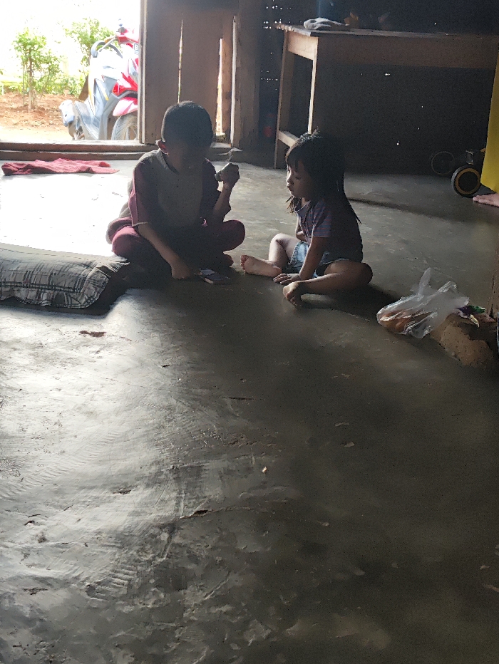 Two young children are seated on a simple, worn concrete or packed earth floor inside a dwelling in Selagai Lingga, Indonesia, during daytime. Natural light streams into the room through an open doorway in the background.
On the left, a boy, dressed in a dark-colored top with lighter sleeves and dark trousers, sits cross-legged. His face is obscured by shadow, but he appears to be holding something small in his right hand, possibly a piece of food or a small object, and looks down. A plaid pillow rests beside him on the left.
To the right, a girl, wearing a light-colored top and shorts, sits with her knees bent, facing towards the boy. Her head is tilted slightly, and she looks towards him. Her bare feet are visible on the floor.
In the middle ground, near the girl, there's a clear plastic bag containing what appears to be snacks or food items. The background reveals a rustic interior with wooden elements. To the right, part of a wooden table is visible, with some items placed on it, and what looks like a child's toy wheel on the floor further right.
Beyond the open doorway, an outdoor area is visible with greenery and what appears to be a red and grey motorcycle parked. The light outside suggests a sunny day. No visible text is present in the image.
