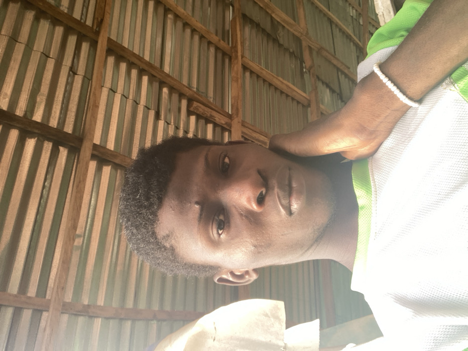 An image depicts a young adult male, likely in Agege, Nigeria, observed from a slightly low angle. The subject, with dark skin and short dark hair, faces forward, looking directly into the camera. A small scar or mark is visible on his forehead above his right eye. He wears a white, mesh-like t-shirt with a light green collar and green accents on the shoulders/sleeves. His left hand is raised, palm against his left cheek and jawline, with fingers slightly splayed. A white beaded bracelet is present on his left wrist. The background features an overhead roof structure composed of wooden rafters supporting corrugated metal sheets, some appearing painted green, creating a striped pattern against unpainted sections. Strong, bright light, originating from the upper left, illuminates the scene, causing glare on the subject's shirt.