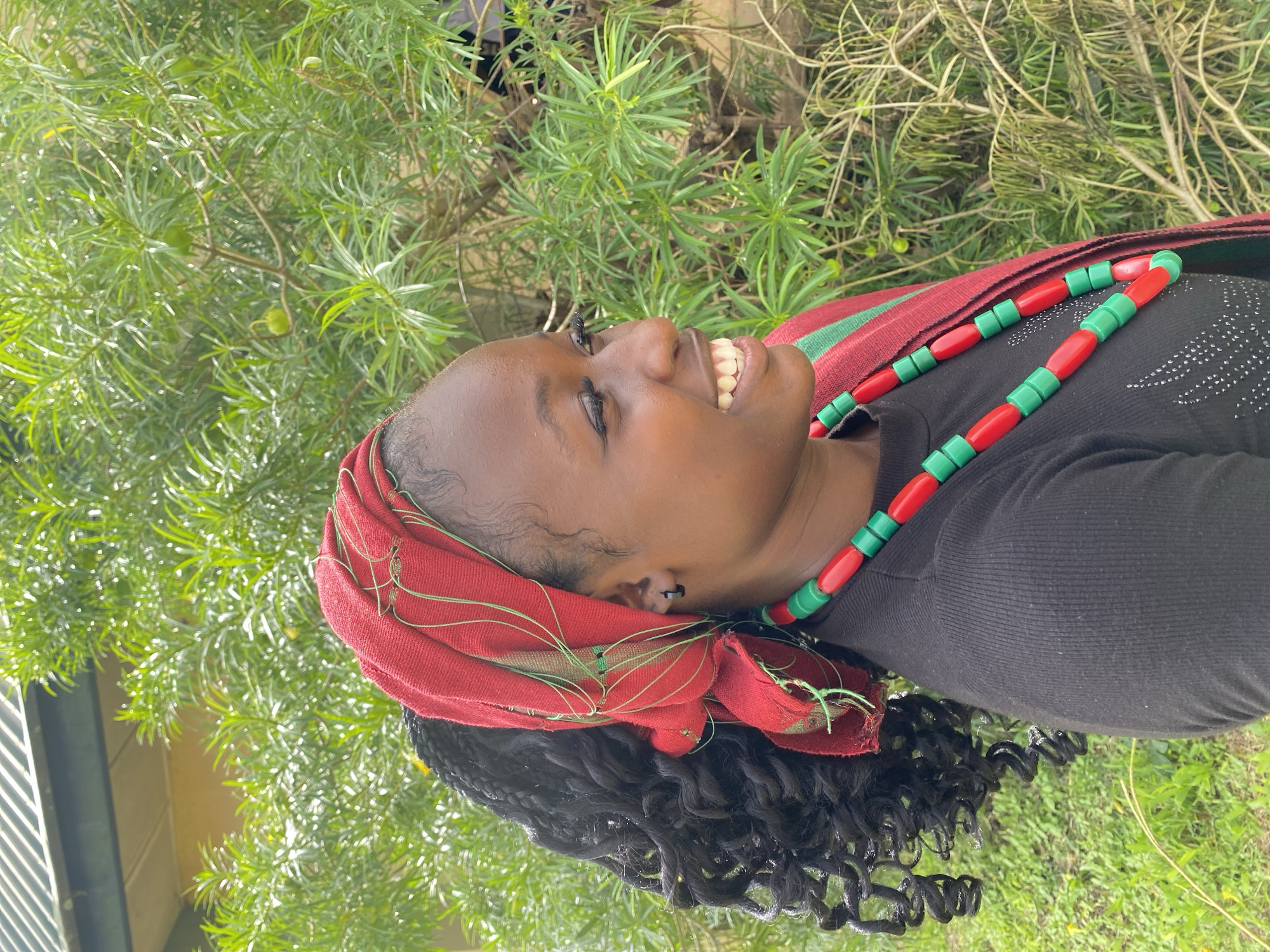 The image shows a young Black woman from the chest up, outdoors. She is wearing a black top, a red and green fabric headscarf, and a necklace made of red and green beads. Her head is tilted back, and she is smiling broadly with her eyes closed. Her hair is dark and curly. The background is filled with lush green foliage, suggesting a natural or garden setting. The lighting indicates it is daytime, likely sunny, with dappled light filtering through the leaves. The woman's expression conveys happiness and contentment. There is no visible text in the image. The location is identified as Wang, Nigeria.