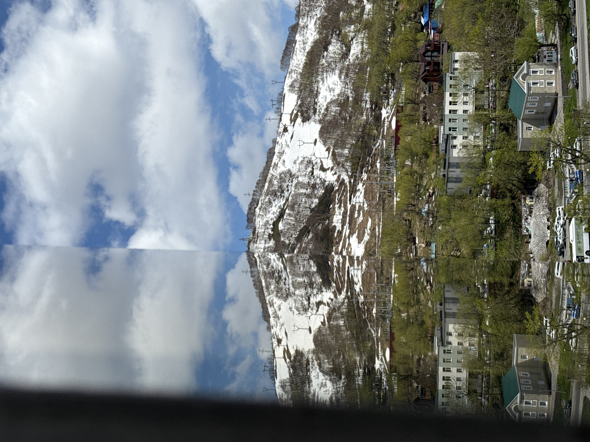 The image displays a mountainous landscape and an adjacent town, captured with a prominent vertical reflection, possibly in still water or a large glass surface. The original scene, which appears in the upper half of the image (if viewed upright after rotating the provided image counter-clockwise by 90 degrees), features a snow-covered mountain with scattered dark, bare trees and patches of green-leaved trees at its base. Several ski lift towers or power lines are visible ascending the mountain slopes, indicating a ski resort or an area with significant infrastructure.
To the right of the mountain's base, a small town or settlement is visible, comprising various multi-story buildings. Some buildings appear residential, while others might be commercial establishments like hotels or apartments. A prominent building with a distinctive green roof stands out. A road with parked cars runs through the town. The sky above is a clear blue adorned with fluffy white clouds.
The lower half of the image is a sharp, almost perfect reflection of the upper scene, mirroring the sky, clouds, mountain, snow, trees, buildings, and vehicles. This reflection creates a strong sense of symmetry and artistic composition. A dark, indistinct band runs across the very bottom of the entire image, likely the edge of a window frame or railing from the vantage point of the photographer.
The scene is set during the daytime under clear weather conditions, with bright sunlight illuminating the landscape. The mix of snow on the mountain and green foliage on trees suggests a transitional season, such as late spring or early summer. While the location is Petropavlovsk-Kamchatsky, Russia, this information is external and not explicitly visible in the image itself, though the mountainous terrain is consistent with the region. No clearly legible text is visible within the image.