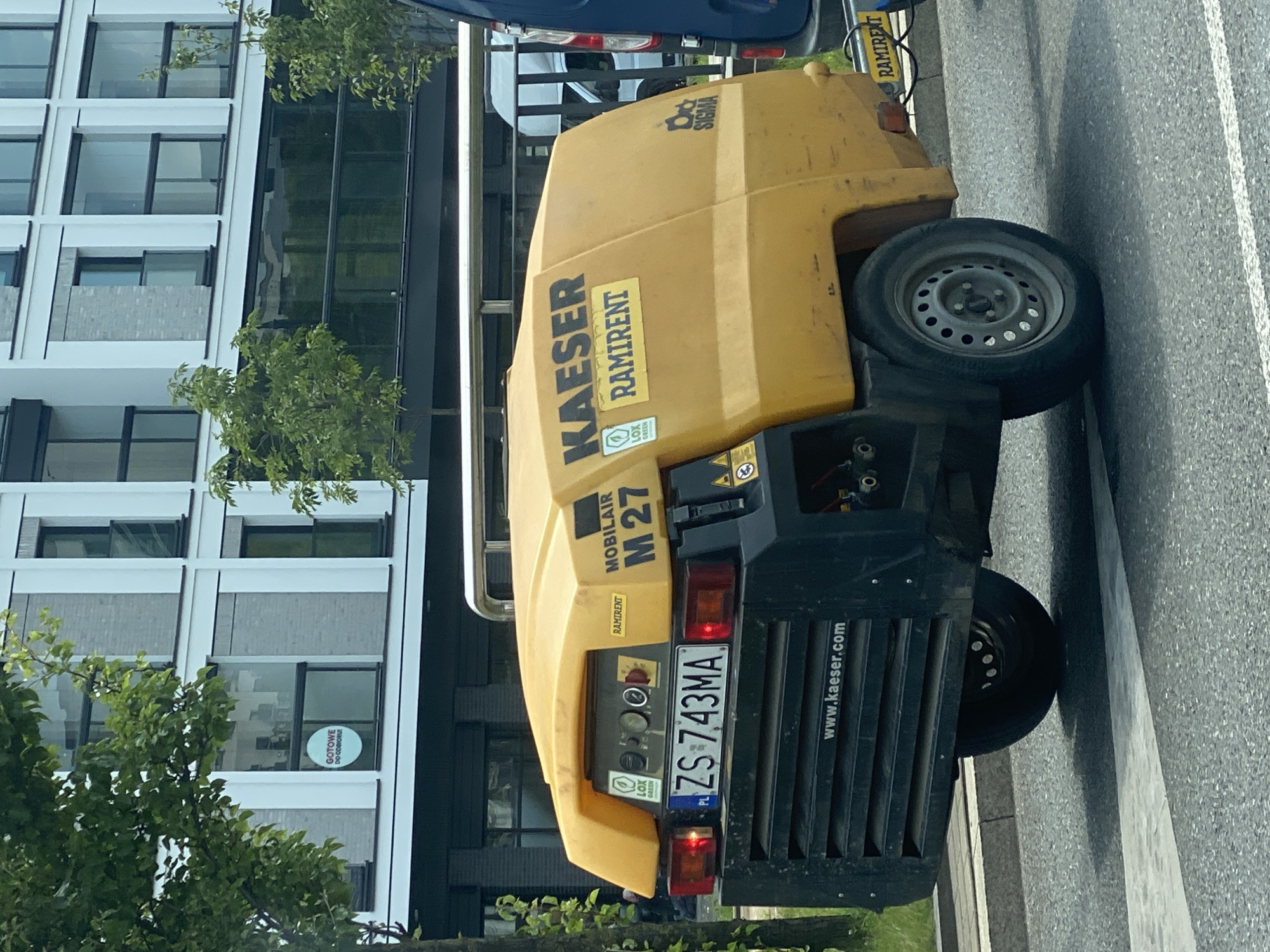 This media file captures a yellow KAESER MOBILAIR M 27 compressor parked on a street in Łódź, Poland. The compressor, a significant piece of industrial equipment, is the primary subject. Its branding is clearly visible, featuring "KAESER" and "RAMIRENT" multiple times. A license plate with the characters "ZS 743MA" is affixed to the rear.
The compressor is positioned next to a modern building with a facade of white panels and large glass windows, some of which reflect the surrounding greenery. Trees with lush foliage are present in the foreground, suggesting a pleasant time of year, possibly spring or summer. A circular sticker on one of the building's windows reads "GOTOWE DO ODBIORU," which translates to "Ready for pickup" in Polish, implying the building might be a commercial establishment or rental service.
The scene appears to be outdoors, and the lighting suggests it is daytime, with no strong indications of specific weather conditions. The street surface is asphalt, marked with white lines, characteristic of a road or parking area. The overall impression is one of a functional, urban environment where industrial equipment is being stored or deployed. The presence of the compressor and the building hints at construction, maintenance, or a commercial rental operation in the vicinity. The image is oriented vertically, with the compressor tilted.