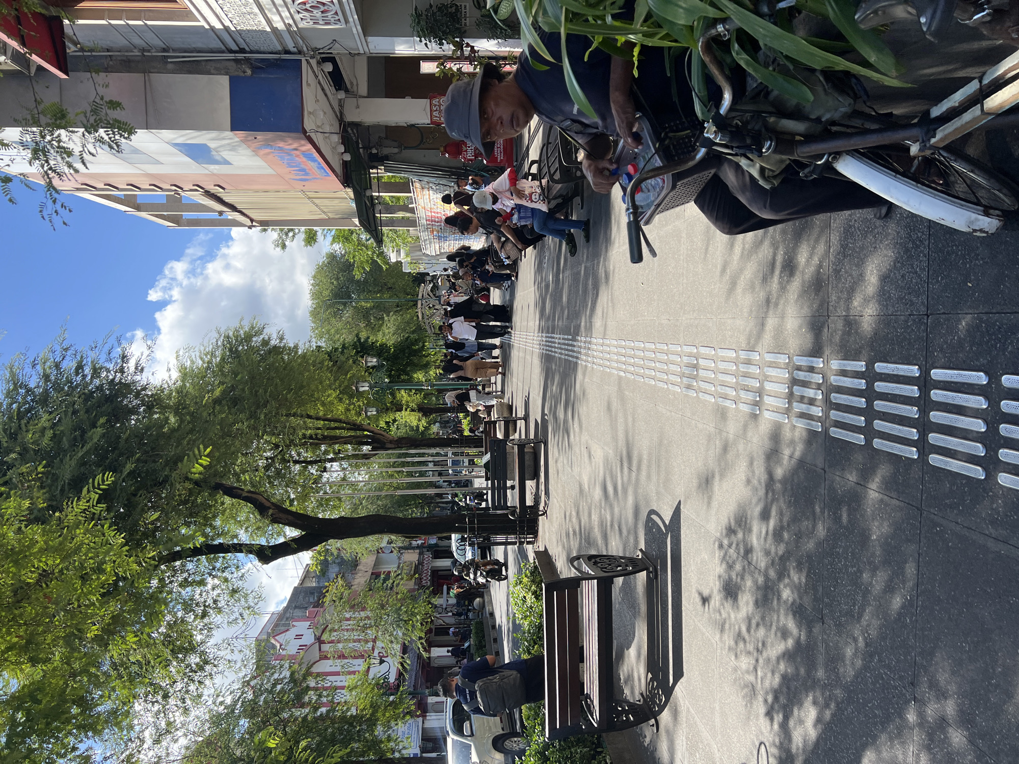 This media file captures a bustling street scene in Yogyakarta, Indonesia, likely during the daytime given the strong sunlight and shadows. The perspective is from a low angle, looking up along a pedestrian walkway lined with trees.
The primary subjects are the people walking and sitting along the sidewalk, interspersed with parked motorcycles and bicycles. A man wearing a hat and dark shirt is prominently featured in the foreground, looking towards the camera while tending to a bicycle. Further down the walkway, several individuals are seated on benches, some with backpacks, suggesting a leisurely stop or waiting period. The architecture of the buildings lining the street is visible, with some showing signs of age and others with colorful facade details. Lush green trees cast dappled shadows on the pavement, creating a pleasant, shaded atmosphere.
The activity taking place is the normal flow of pedestrian and vehicular traffic in a public urban space. People are moving about their day, suggesting a busy marketplace or tourist area. The presence of benches indicates a place for rest and observation.
Notable details include the textured paving stones on the walkway, which appear to be tactile paving for the visually impaired. The sunlight is bright, indicating a clear or partly cloudy day. The overall mood seems relaxed and ordinary, reflecting a typical day in this Indonesian city.
Some visible text can be seen on the building facades, though it is partially obscured and difficult to read in full. One sign appears to have red lettering, possibly indicating a shop or establishment. Another sign reads "JASA" which translates to "SERVICE" in Indonesian. The image provides a glimpse into the vibrant street life and urban environment of Yogyakarta.