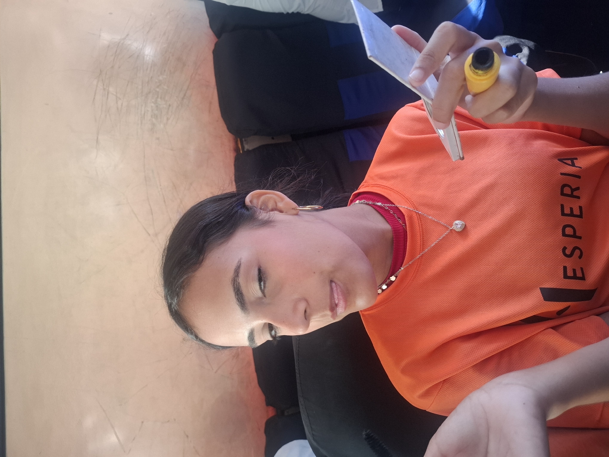 A person is seated in an indoor environment, wearing an orange t-shirt with the word "ESPERIA" printed on it. The person is holding a yellow-handled instrument and a white card. They have dark hair and are wearing a silver necklace. The setting appears to be indoors, and the background is a light orange wall. There is no information in the image to indicate it is in Juan Griego, Venezuela.