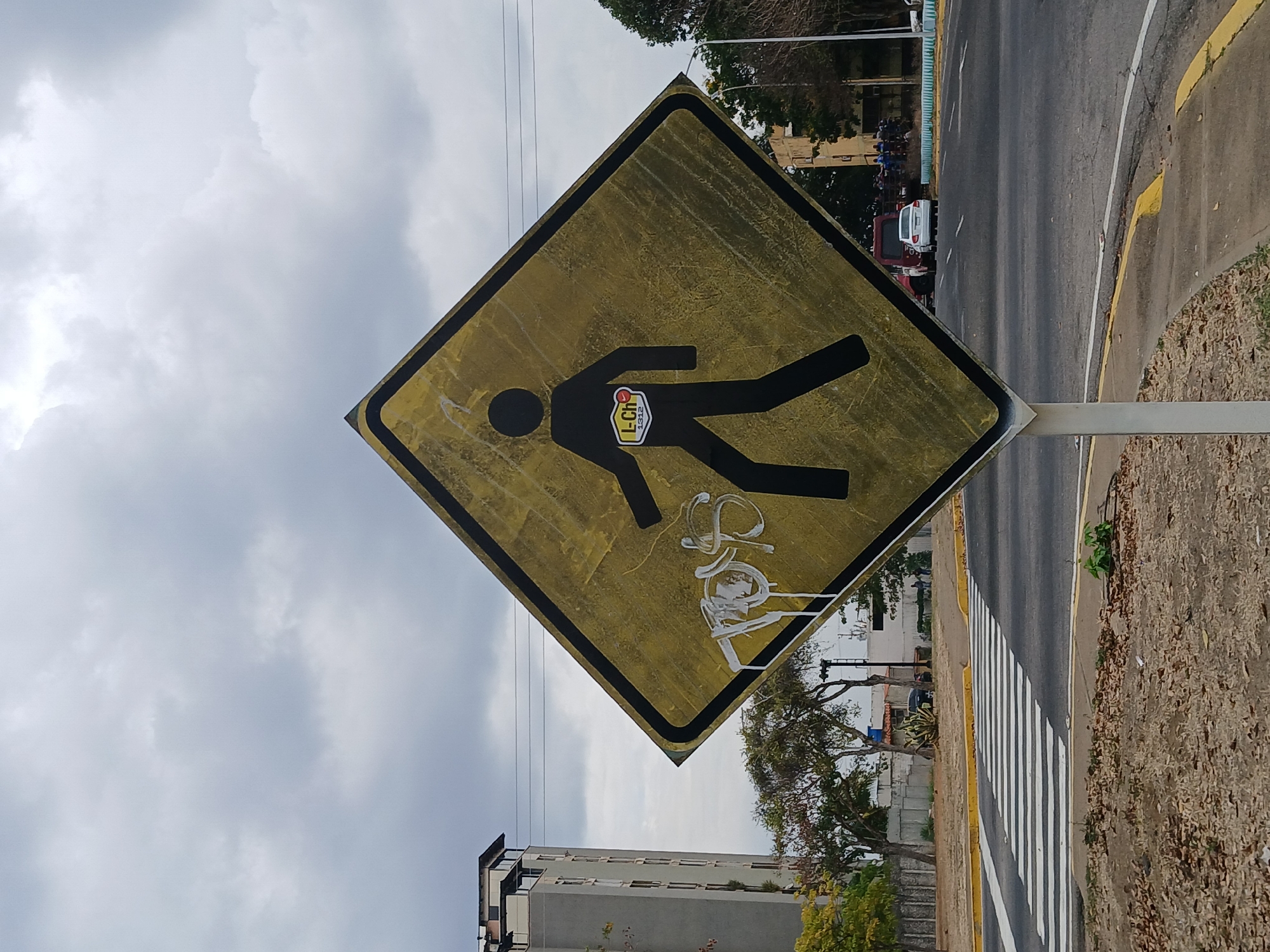 A medium shot of a yellow and black pedestrian crossing sign that has been vandalized with white graffiti. The sign is diamond-shaped and has an image of a person walking in black. A sticker is placed on the person's body, and white graffiti is sprayed at the bottom of the sign. The sign is located near a road with a crosswalk. The location is Guayana City, Venezuela. The sky is cloudy.