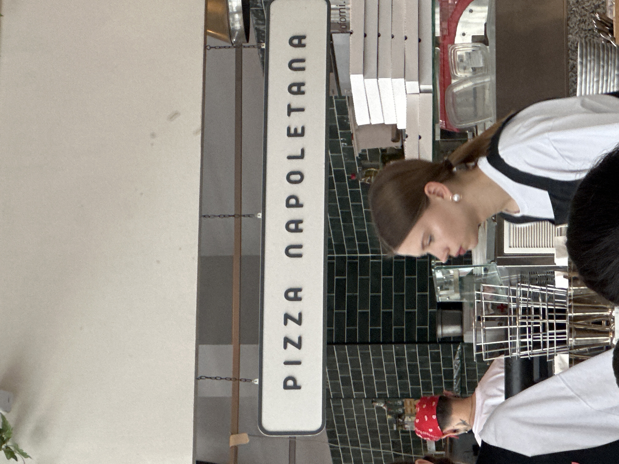 This image captures an indoor scene inside a food establishment, likely a pizzeria, located in Nizhny Novgorod, Russia. The perspective is from the customer's side, looking towards a service area where staff are at work.
A prominent, long, rectangular white sign hangs vertically on the left side of the frame, suspended by chains. The sign displays the black capital letters: "PIZZA NAPOLETANA."
Behind the counter, two individuals are visible. On the right, a woman with brown hair tied back in a ponytail, wearing a white shirt with black detailing and a pearl earring, is leaning forward with her head down, focused on an activity at the counter. Below and slightly to her right, another person, wearing a red bandana and a white shirt with black accents, is partially visible from the back, also engaged in tasks behind the counter.
The background features a wall covered in dark green or black subway-style tiles. Above these tiles, various kitchen or service items are present, including metal racks, clear containers, and a white ventilation grate. Faint text reading "domi." can be seen on what appears to be a stack of boxes or papers behind the counter. The left side of the image shows a plain, light-colored wall. The scene depicts a work environment within a food service setting.