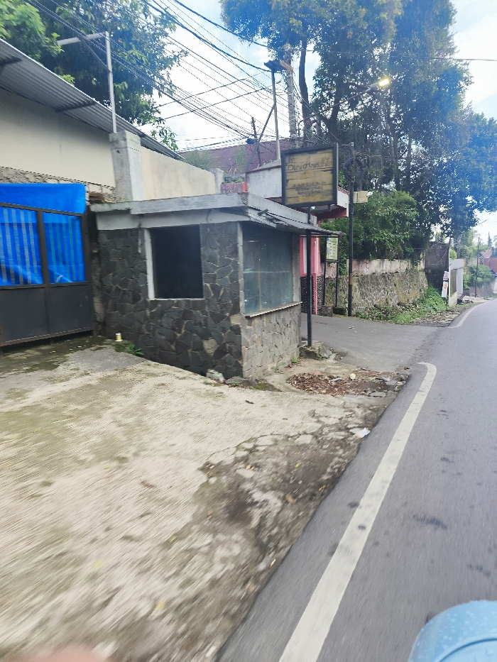 The image shows a view from a car driving down a paved road in Lembang, Indonesia, with coordinates -6.82 latitude and 107.611 longitude. The road has a white dividing line. On the right side of the road, there is a stone building with a glass window and a sign above it with the text "Dendrologi". The building appears to be a guardhouse or a small shop. There is a stone wall behind the building, with green foliage growing along its top. On the left side of the road, there is a concrete driveway with a blue metal gate. The gate is partially closed and the area appears to be a private residence. In the background, there are trees and power lines. The weather is cloudy and the sky is a light blue.