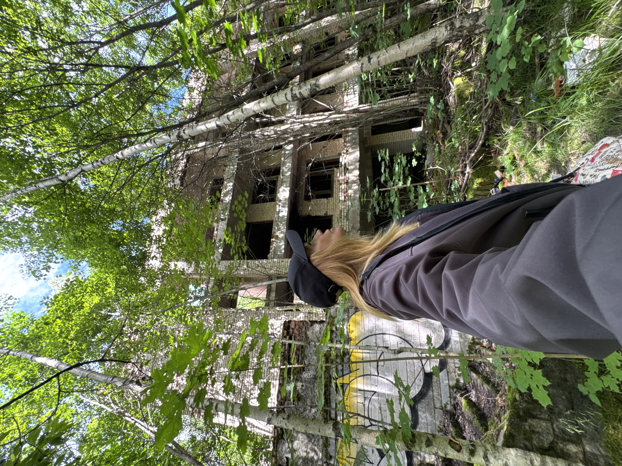 A young woman with blonde hair, wearing a dark baseball cap and a dark jacket over a patterned shirt, stands in a forest looking up at an abandoned brick building. The building is multi-story with visible broken windows and graffiti on the lower levels. Trees, some with birch bark, grow around and through the structure, with dense green foliage obscuring parts of the building and the sky. The ground is covered in leaves, grass, and some scattered debris. The location is the city of Interkompleks, Russia.