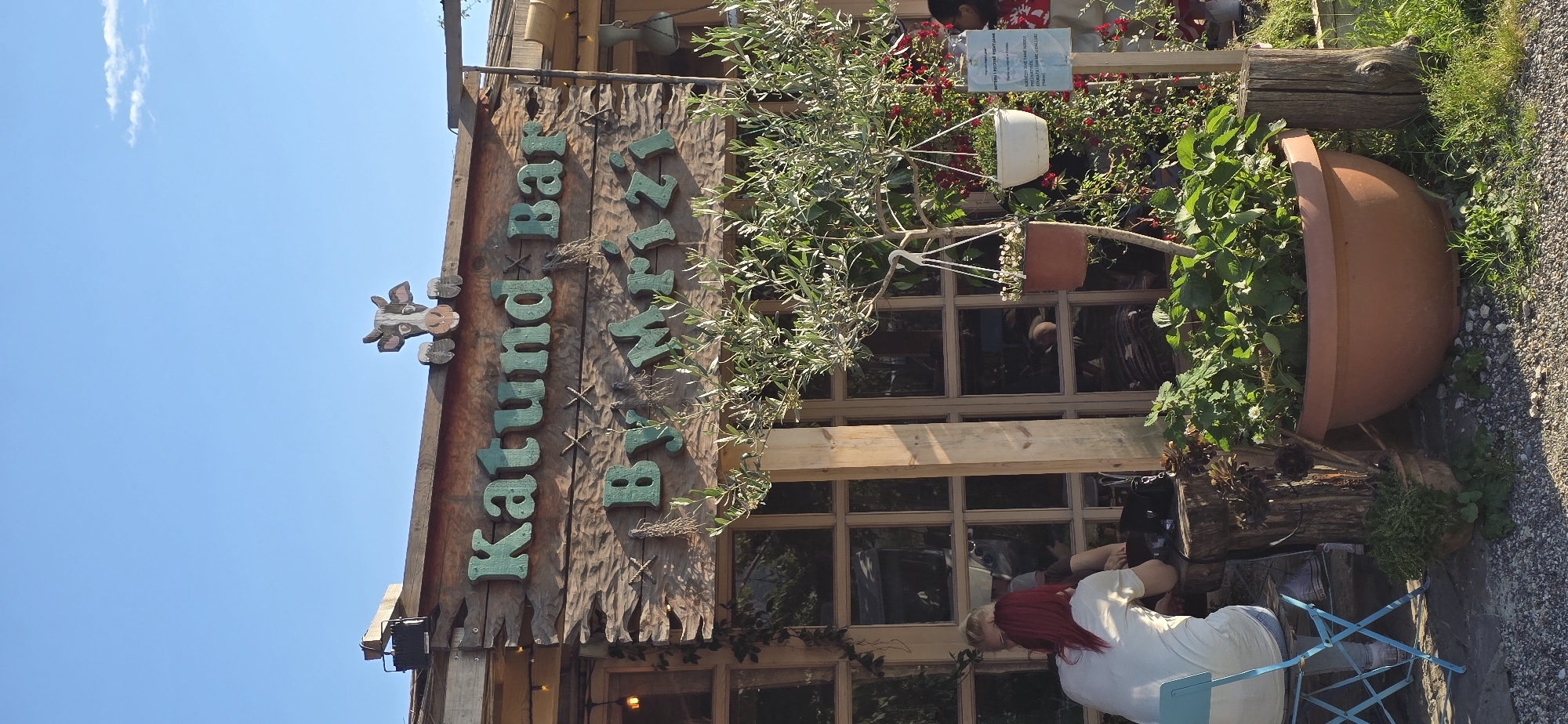Captured at an angle against a clear, blue sky, the scene features a building in Blinisht, Albania, identified by a sign that reads "Katunda Bar by Wirtz." The building's facade is adorned with various plants and decorative elements.
A person with red hair is seated on a blue chair near the entrance, appearing to be engaged in some activity. The surroundings include a wooden structure with windows, potted plants, and a large earthen pot with foliage. The ground is covered with gravel or small stones.