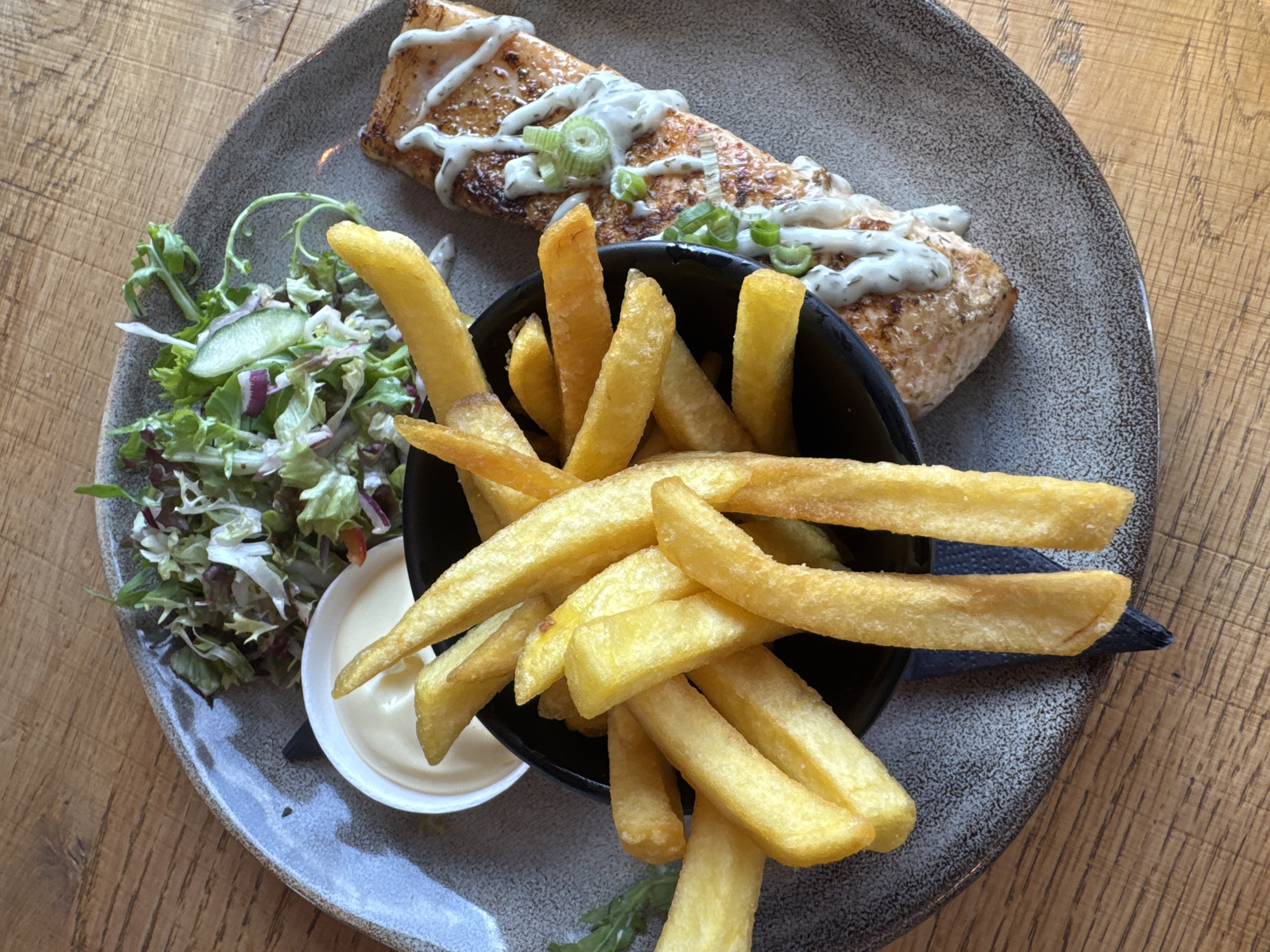An overhead shot displays a single, oval-shaped ceramic plate with a speckled grey-brown finish, positioned on a light brown wooden table featuring visible grain. The plate holds a meal consisting of three primary components. To the right, a rectangular piece of grilled salmon, light orange-pink in color, is drizzled with a thick, white, creamy sauce and garnished with chopped green scallions. In the center, a small, dark black bowl is overflowing with golden-brown french fries, some spilling out onto the plate. To the upper left, a small, white ramekin contains a creamy white condiment. Adjacent to the ramekin and above the salmon, a pile of mixed green salad, featuring various lettuce leaves, some red cabbage, and thinly sliced cucumber, completes the meal. A dark blue napkin is partially visible beneath the french fries. No people are present in the frame.