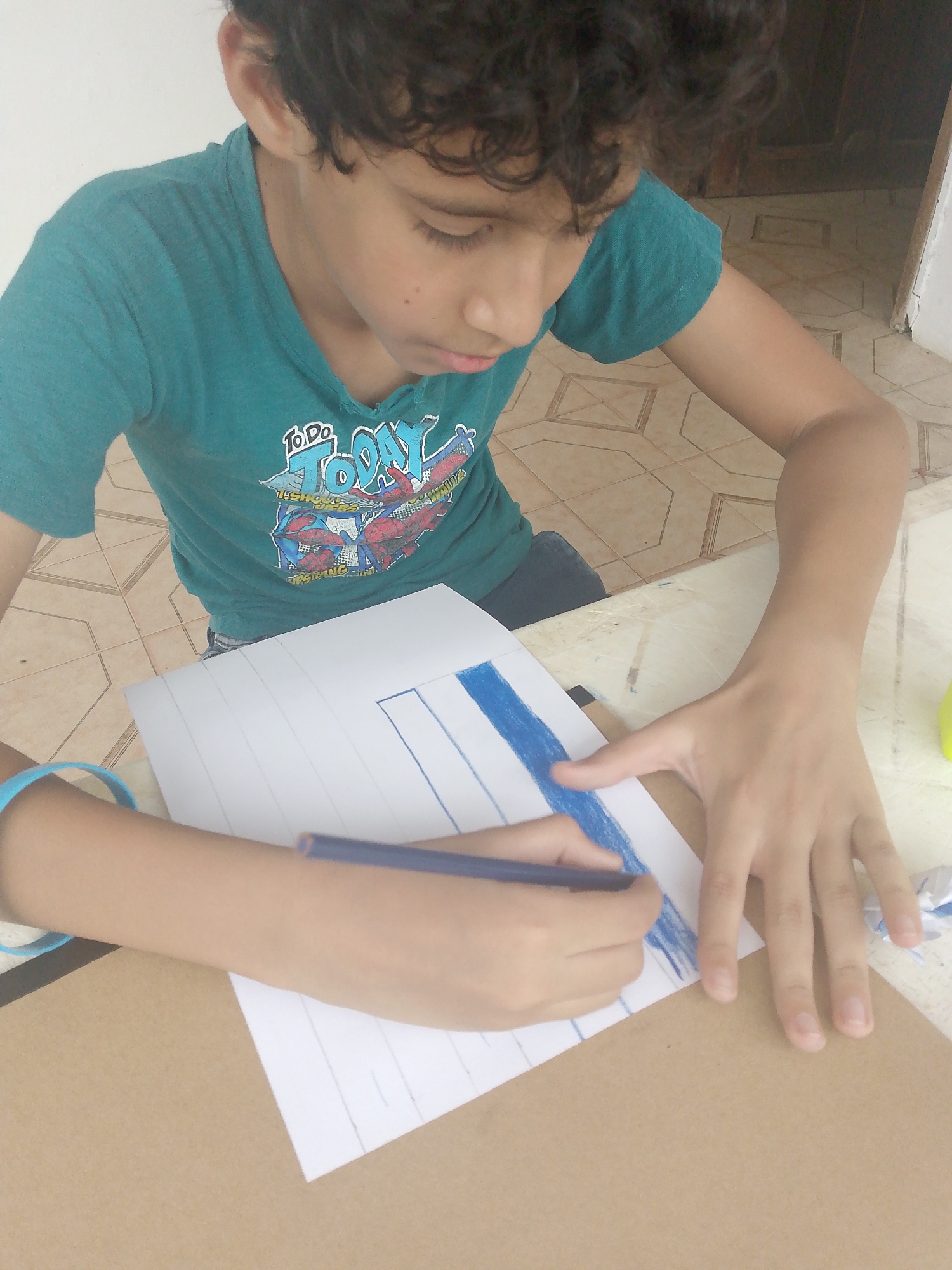 A young boy with curly dark hair, wearing a teal t-shirt with a graphic on it, is drawing with a blue colored pencil on a piece of white paper that has vertical lines on it. He is seated at a table, part of which is covered with brown cardboard. The setting is inside a building with light-colored tile flooring, and a door is visible in the background. The image was taken in Tinaquillo, Venezuela.