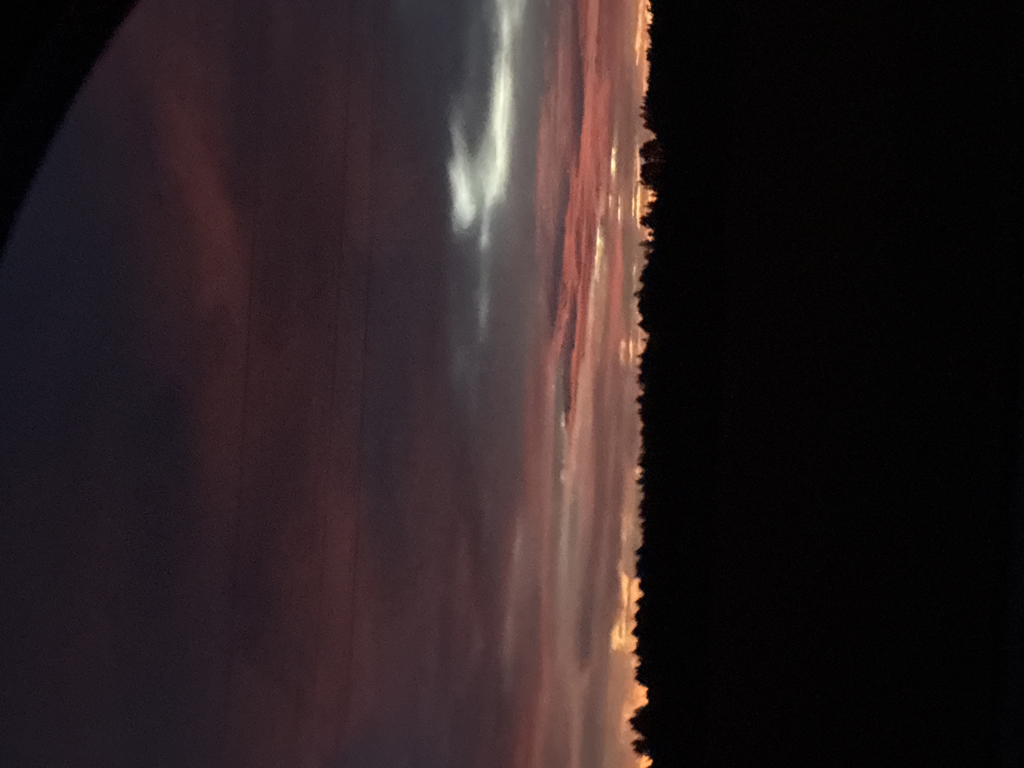 The image depicts a dusky sky with wispy clouds, tinted with shades of pink and orange, suggesting either sunrise or sunset. The right edge of the frame is dominated by a dark, silhouetted treeline against the colorful sky. The majority of the image is composed of the sky, with a hint of darker, possibly purplish clouds on the left. The overall mood is serene and natural, indicative of the transition between day and night. There is no visible text, people, or distinct objects other than the natural elements of the sky and trees. Given the location context of Kotelnich, Russia, this could be a typical rural or natural landscape view.