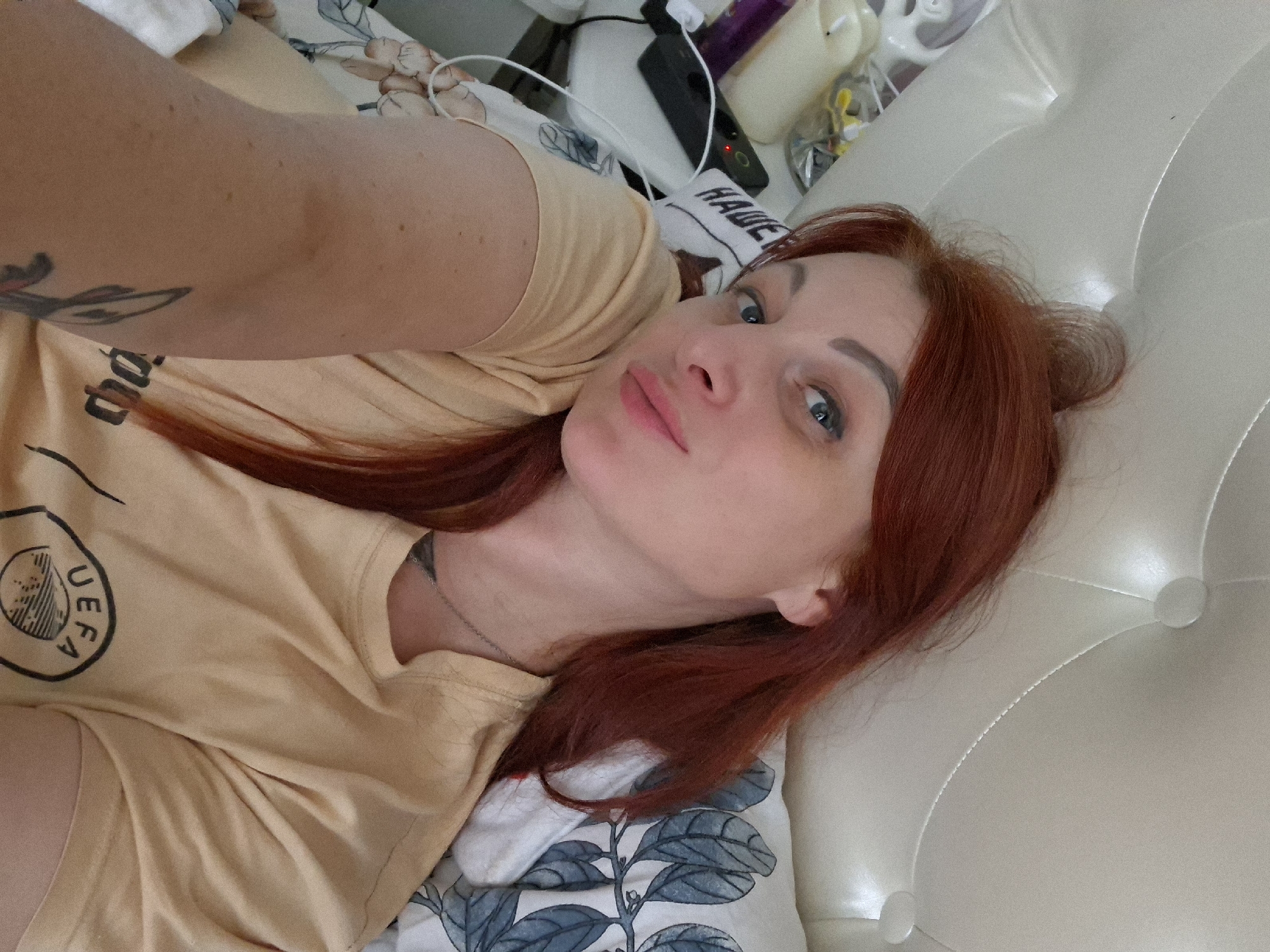 A selfie of a woman with red hair lying in bed. The woman is wearing a yellow t-shirt with a UEFA logo on it. She has blue eyes and pale skin. There are pillows with a leaf pattern and a white tufted headboard in the background. A power strip and a phone charger are visible on a nightstand next to the bed. The scene appears to be indoors, in a bedroom. The location is not specified in Myadzyel, Belarus.