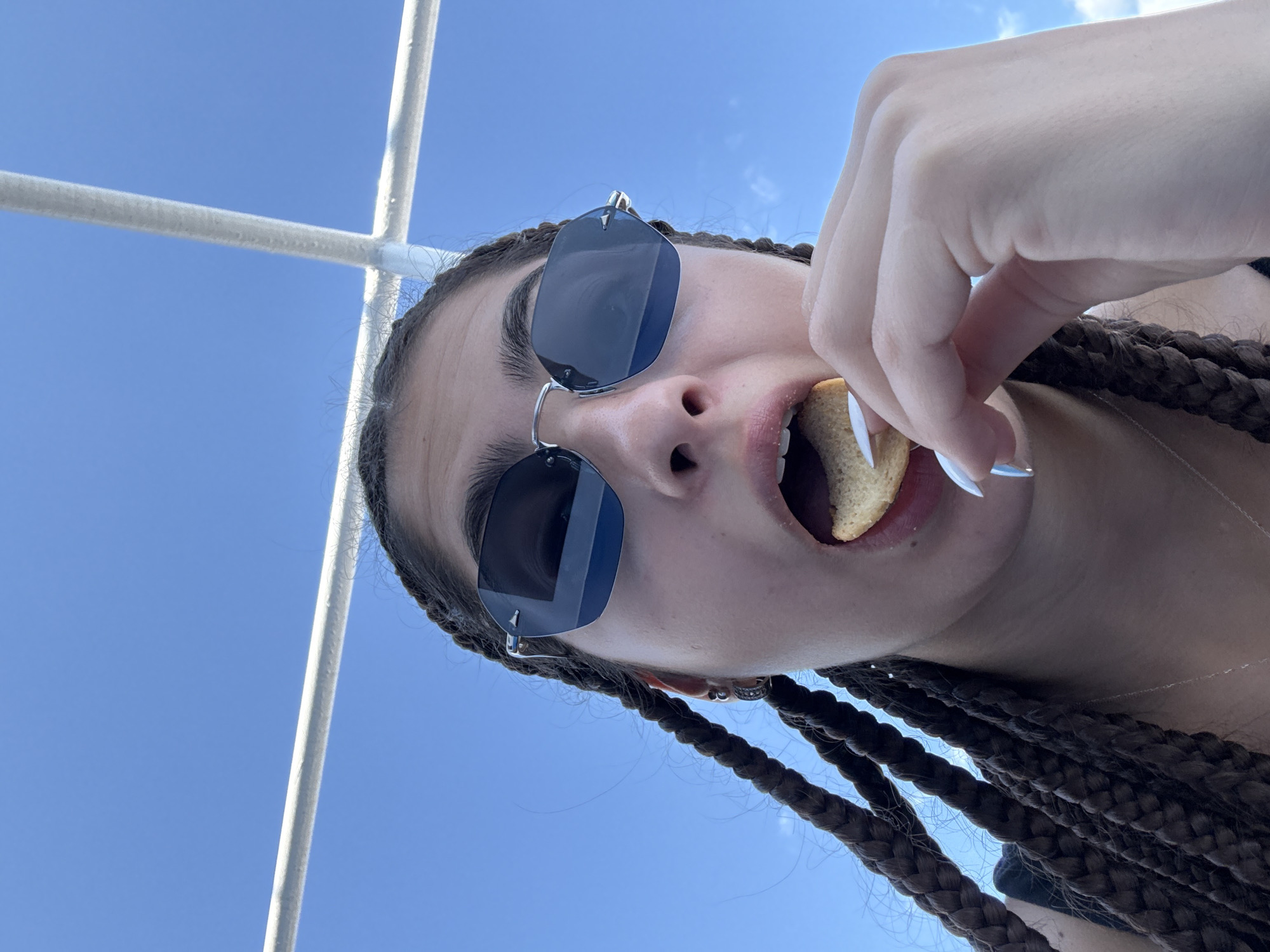 A close-up, low-angle, outdoor, selfie-style photo shows a young person with dark braided hair wearing large, dark-lensed sunglasses. They are holding a light-colored cracker to their open mouth with their right hand, appearing to be eating it. The person is outdoors with a clear blue sky and white structures, possibly part of a boat or a canopy, visible in the background. The location is specified as Prinos, Greece.