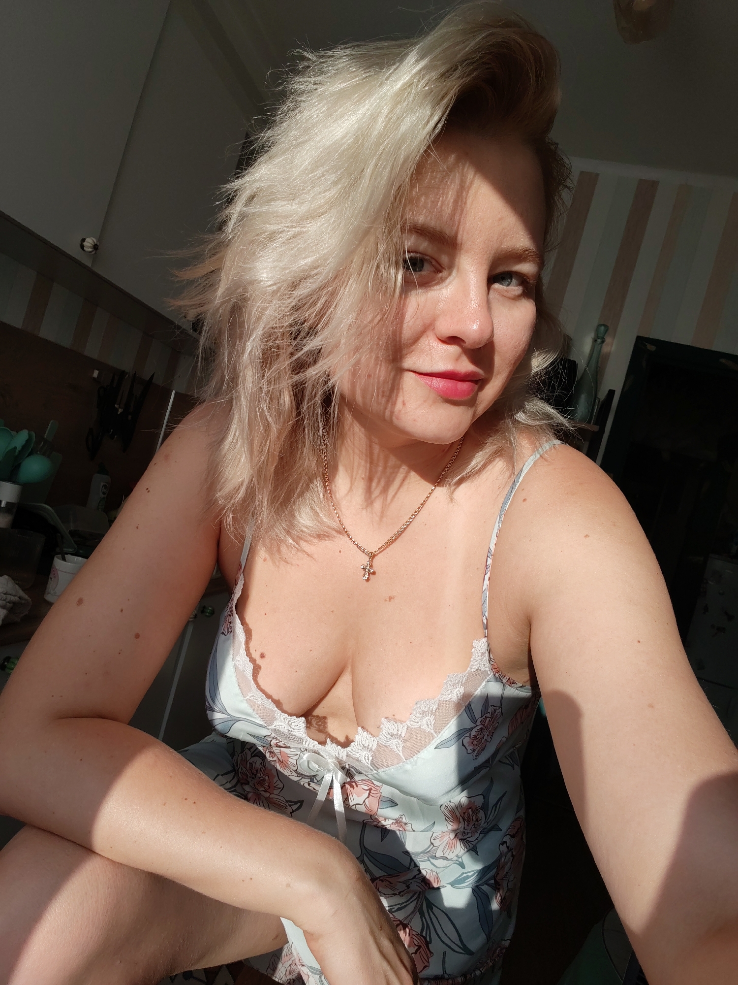 The image shows a woman with blonde hair and blue eyes, wearing a light blue nightgown with a floral print and a gold cross necklace. She is sitting and looking at the camera, with sunlight casting shadows on her face and body. The background appears to be an indoor setting, possibly a home, with some kitchen items visible. There is no information to indicate the specific location within the city of Izhevsk, Russia, or any particular activity taking place.