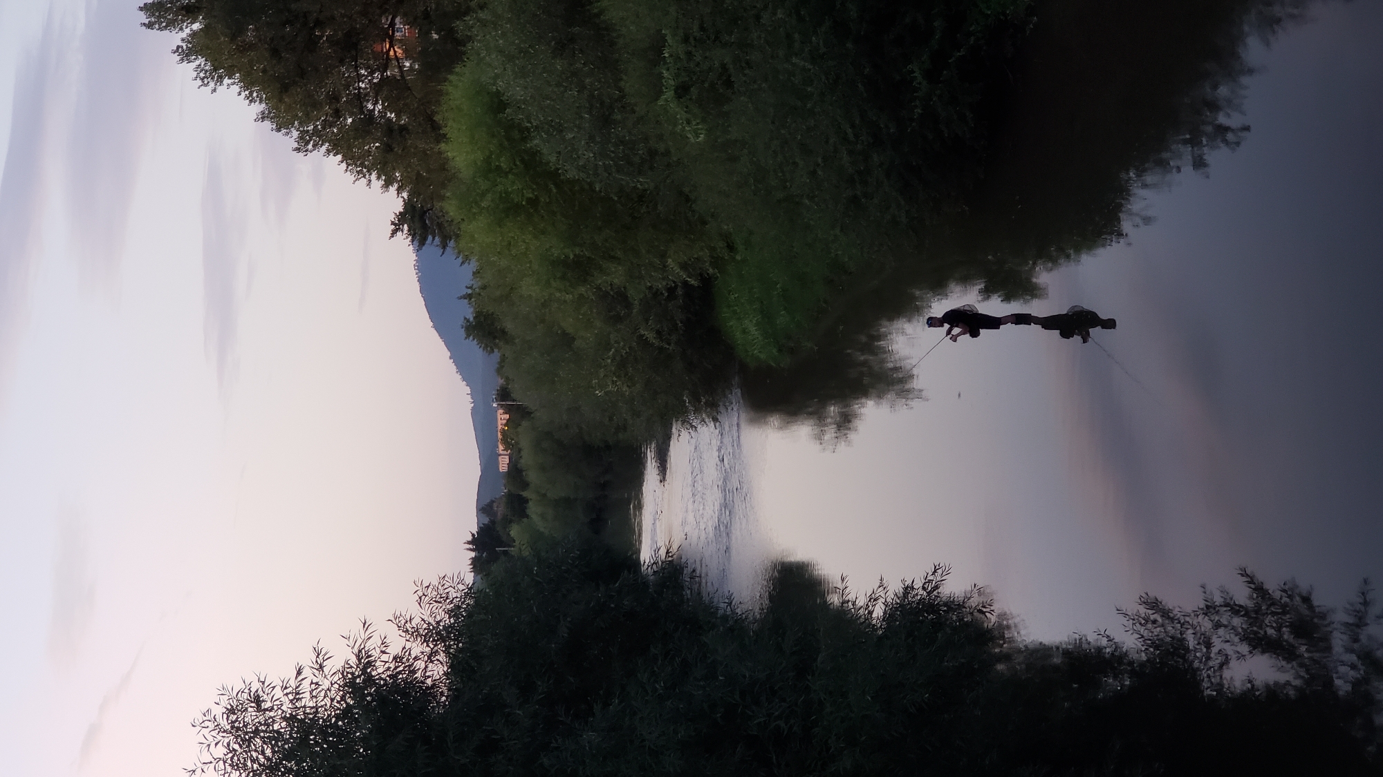 A single male is standing in the middle of a wide river, fishing. He is silhouetted against the soft, twilight sky, holding a fishing rod. His reflection is clearly visible in the calm water. The riverbanks are lined with lush, dark green trees, which cast shadows into the water. In the distance, hills and some buildings are visible under a pale purple and grey sky. The scene appears to be a peaceful, natural setting. This could be the Torysa River in Prešov, Slovakia, known for its natural beauty and recreational opportunities.