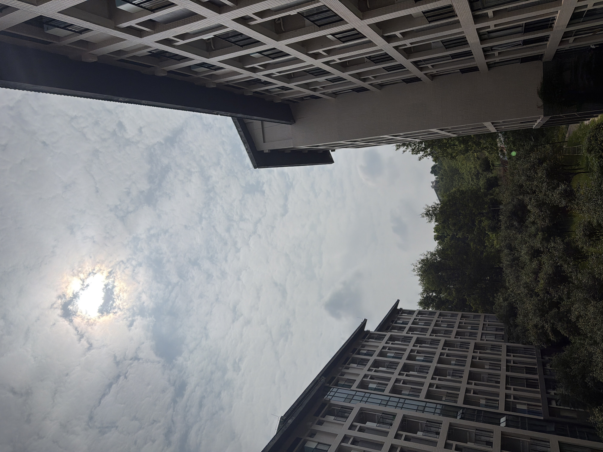 The image shows an outdoor, daytime scene with two modern buildings and a lush green hillside covered in trees. The sky is cloudy with the sun visible through a break in the clouds. The buildings appear to be residential or dormitory-style, with multiple stories and rows of windows and balconies. The building on the right is closer and angled towards the viewer, while the building on the left is further away and also angled. The hillside behind the buildings is densely vegetated, suggesting a natural or park-like setting. The overall impression is one of a relatively peaceful and possibly suburban or semi-rural environment within a city. The architecture of the buildings suggests a contemporary design.