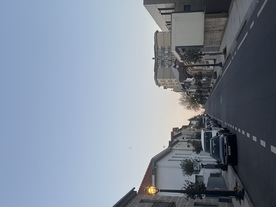 The image depicts an uphill street scene in Guimarães, Portugal, specifically Rua de São Dâmaso, observed during twilight conditions. The sky transitions from a pale blue at the zenith to a light orange-pink hue near the horizon, indicating either sunrise or sunset. No clouds are present.
The street, paved with asphalt, ascends into the distance, with a white dashed line visible on the right side, demarcating the traffic lane from the parked vehicles. Streetlights illuminate portions of the scene with an orange glow; a prominent black street lamp with an illuminated luminaire stands on the left foreground.
On the left side of the street, a line of vehicles is parked parallel to the curb, facing uphill. The closest vehicle is a black Audi sedan, with its license plate "57 GD-186" partially visible. Behind it, a white van and several other indistinct cars are parked. The sidewalk on this side is raised with a stone curb, featuring small, manicured trees planted beside street lamps. Adjacent to the black Audi is a dark parking meter or information post, and a white sign with a blue 'P' for parking is also visible. Buildings lining this side are predominantly light-colored with red-tiled roofs, the closest showing exposed stone elements and a sign that reads "PAPABOA."
The right side of the street features a mixed architectural style, including older buildings with tiled roofs and more modern structures with flat roofs and varying facades, including a section with dark cladding and balconies. Further up the street, a multi-story residential building with a beige/grey facade and numerous windows is visible. Small trees are also planted along this sidewalk. No individuals are present in the frame.