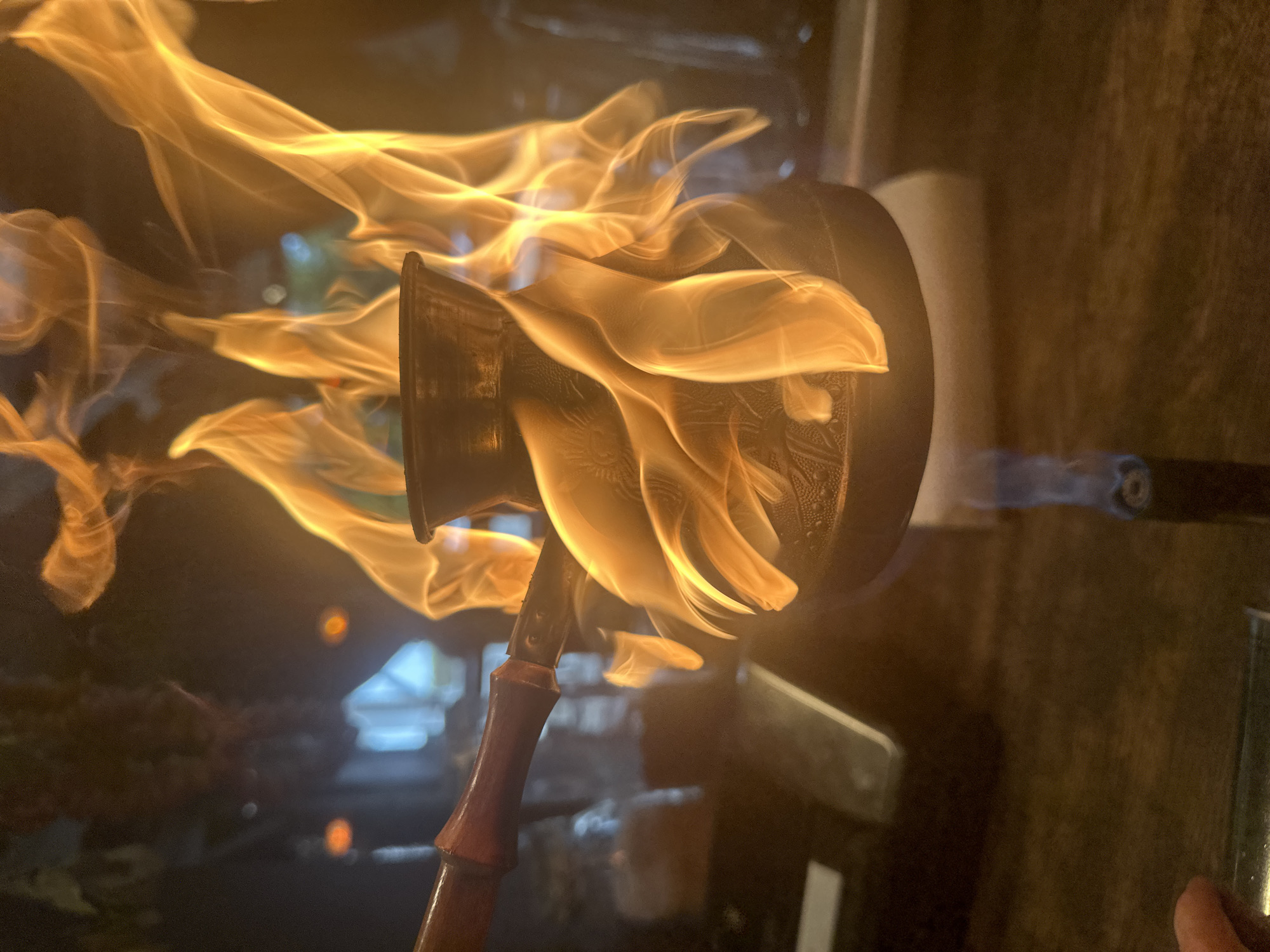 A black ibrik, a Turkish coffee pot, is engulfed in flames. The ibrik has a long, red handle. The flames surround the body of the pot, with the fire appearing to originate from a blowtorch off to the right. The scene is indoors, and the background is dark with some blurred shapes and lights suggesting a kitchen or restaurant setting. The city location is Новоглаголево, Russia.