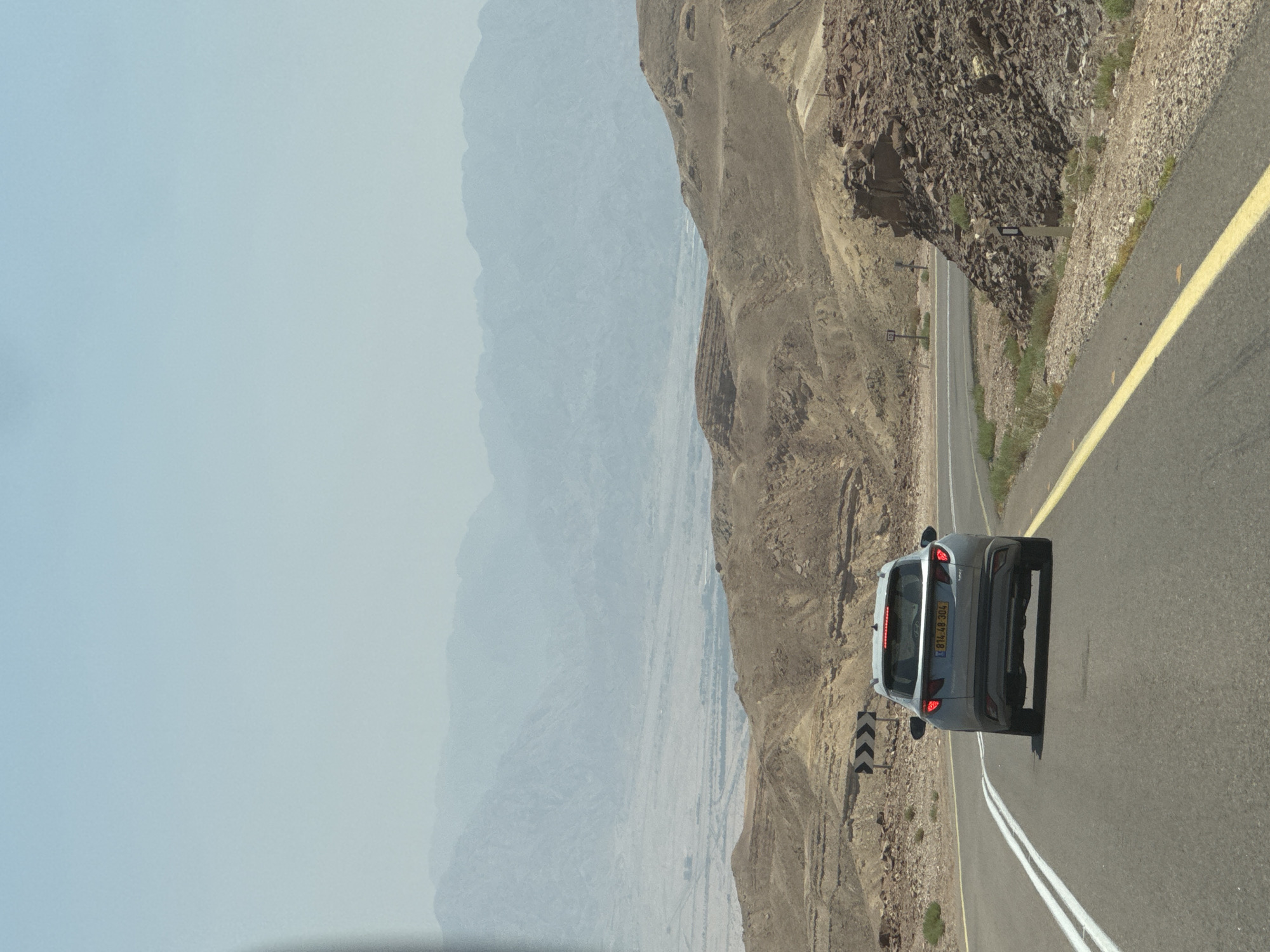 The image captures a view from a moving vehicle on a desert highway, likely near Eilat, Israel, given the arid landscape and mountainous backdrop. A light-colored SUV is driving ahead on the asphalt road, which has a double white line on the left and a single yellow line on the right. The terrain is characterized by rolling, rocky hills with sparse vegetation. In the distance, several layers of hazy mountains are visible, fading into a light blue sky. The sun appears to be high, casting a bright light on the scene. A road sign with chevron markings is visible on the left side of the road. The overall atmosphere suggests a hot, dry environment typical of the region.