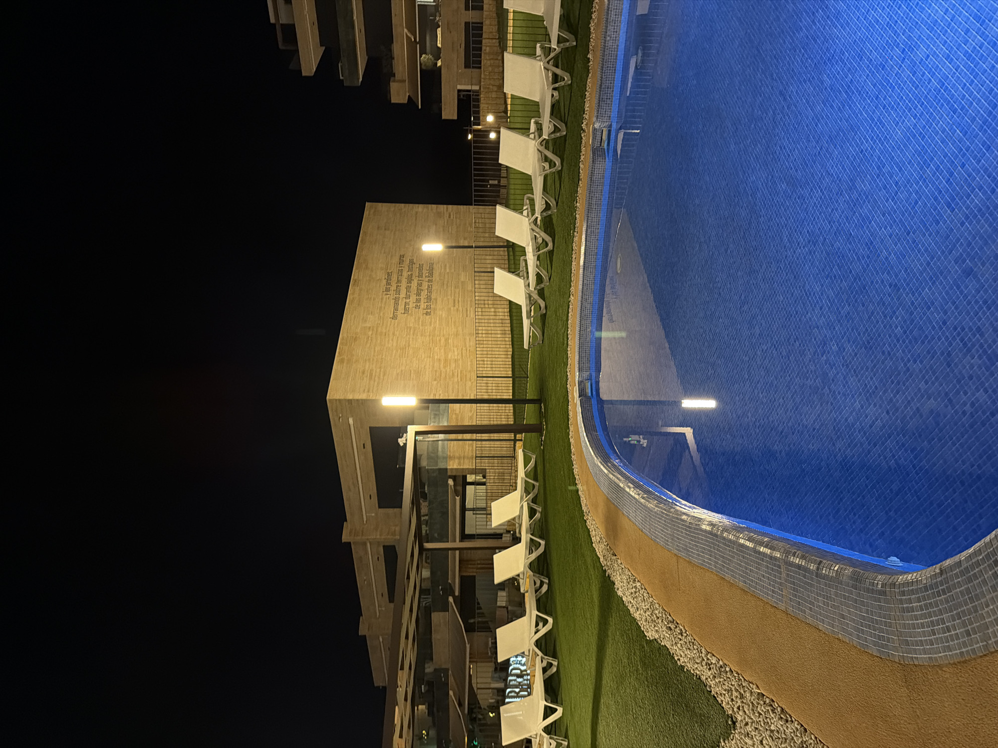 The image depicts an outdoor swimming pool area at night. The pool's water is a vibrant blue, illuminated by overhead lights, creating a serene atmosphere. The pool's edge is lined with mosaic tiles, and beyond that, a strip of tan-colored sand gives way to a lush green lawn. Several white lounge chairs are neatly arranged on the grass, suggesting a place for relaxation.
Adjacent to the pool area is a modern building with a light-colored, textured facade. A prominent feature of the building is a large inscription in Spanish, partially visible and illuminated by a nearby lamppost. The text reads, "y los jardines derramando sobre terrazas y munis fueron durante siglos, testiges de las alegrias y desvelas." This translates to, "and the gardens pouring over terraces and cities were, for centuries, witnesses of joys and sleepless nights." The architecture suggests a contemporary resort or residential complex.
The scene is set at night, indicated by the dark sky and artificial lighting. There are no people visible in the frame, adding to the quiet and tranquil mood. The location is identified as Finestrat, Spain. The overall impression is one of a peaceful, well-maintained leisure space, bathed in the soft glow of night.