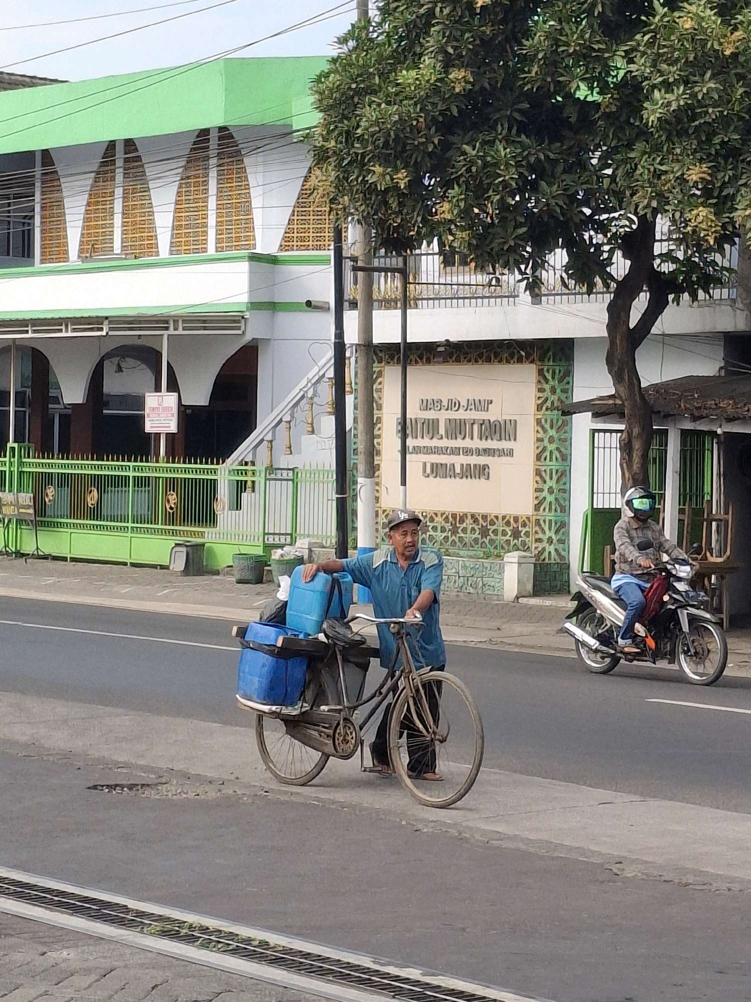 This is a street-level, eye-level, vertical, medium shot photograph taken in Lumajang, Indonesia. In the foreground, a man wearing a blue shirt and a cap stands beside a bicycle. The bicycle is loaded with two large blue plastic containers and other items. The man is looking towards the right side of the frame. To the right of the man, a person wearing a plaid shirt, blue jeans, and a helmet is riding a motorcycle. The motorcycle has a red fender and is moving to the right.
In the background, a building with a green roof and white facade is visible. This building appears to be a mosque, identified by signage that reads "MASJID JAMI' BAITUL MUTTAQIN LUMAJANG". The building features arched windows and a decorative facade. A tall tree with green foliage stands to the right of the mosque. The street in front of the mosque and the building is paved, with a sidewalk separating it from the road. Power lines are visible in the sky above the scene.