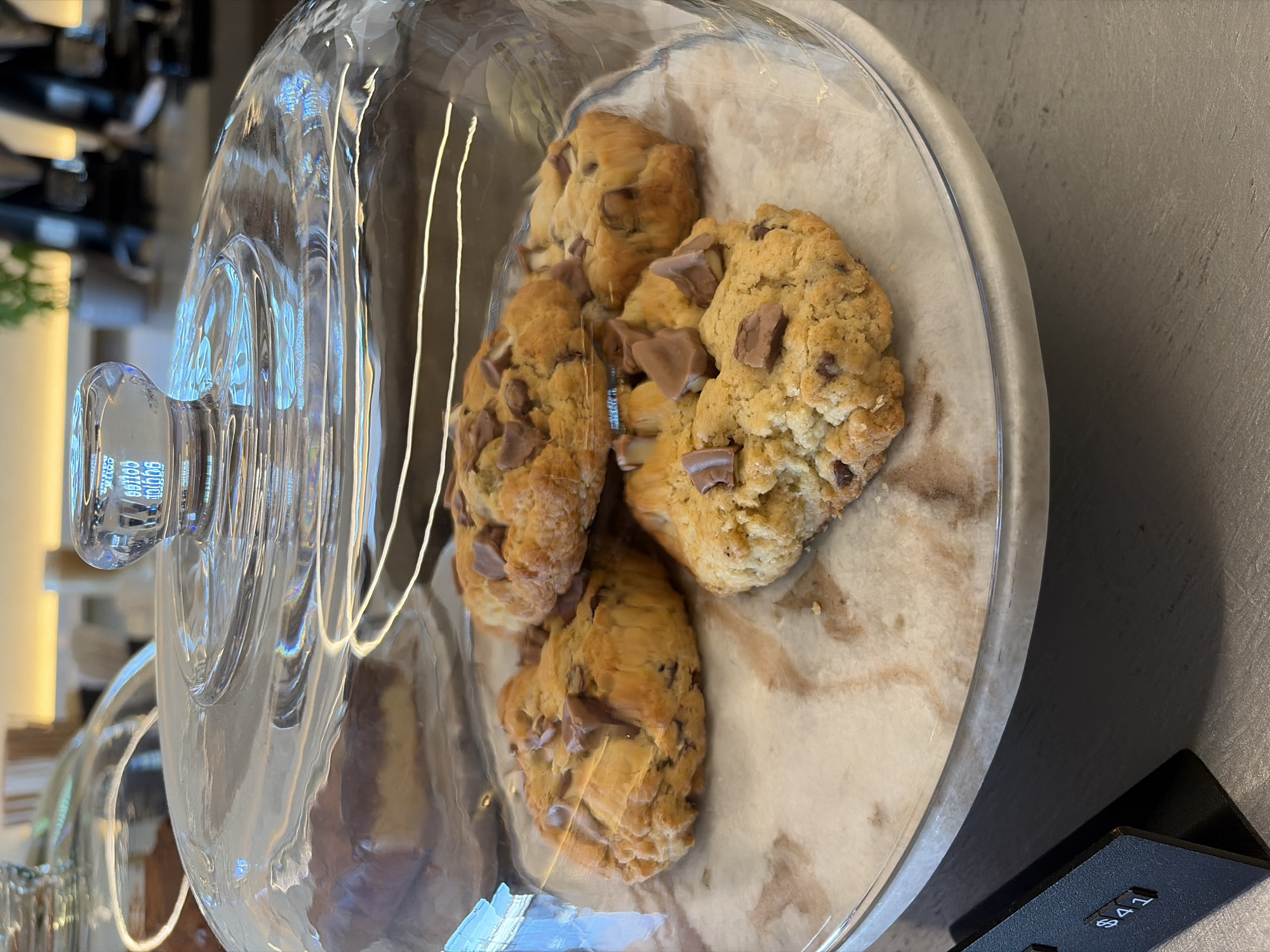 A high-angle, close-up shot depicts a glass domed cake stand, resting on a grey countertop. Inside, on a piece of parchment paper, are four chocolate chip cookies. The cake stand base appears to be marble, and the stand sits beside a small, black rectangular sign with white lettering that reads "641." The background shows a blurred interior, possibly a bakery or café, with visible lighting fixtures and shelving. The location is Unknown, Unknown.