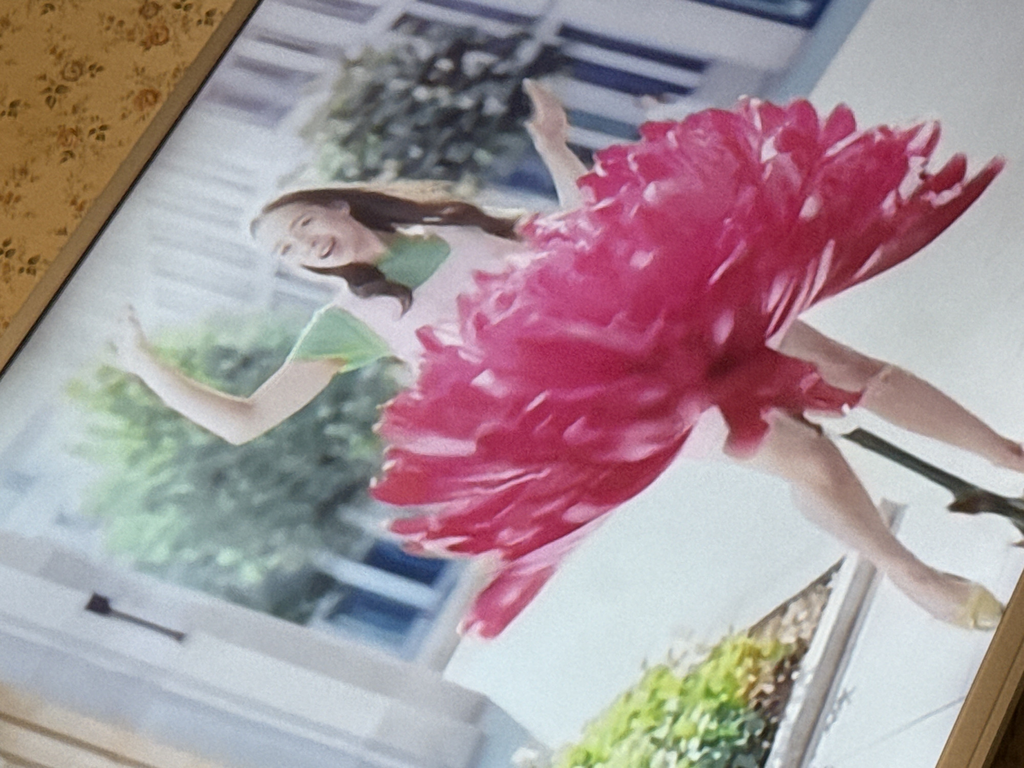 A woman in a pink and white dress is dancing or jumping in the air. The dress has a large, puffy red floral skirt. She has brown hair and is smiling. The background includes foliage and architectural elements like a window. The image appears to be a photograph taken of a screen displaying the scene. The photograph includes visible patterning of the wall on the left side of the screen. No information about the location within Mytishchi, Russia can be determined from this image.