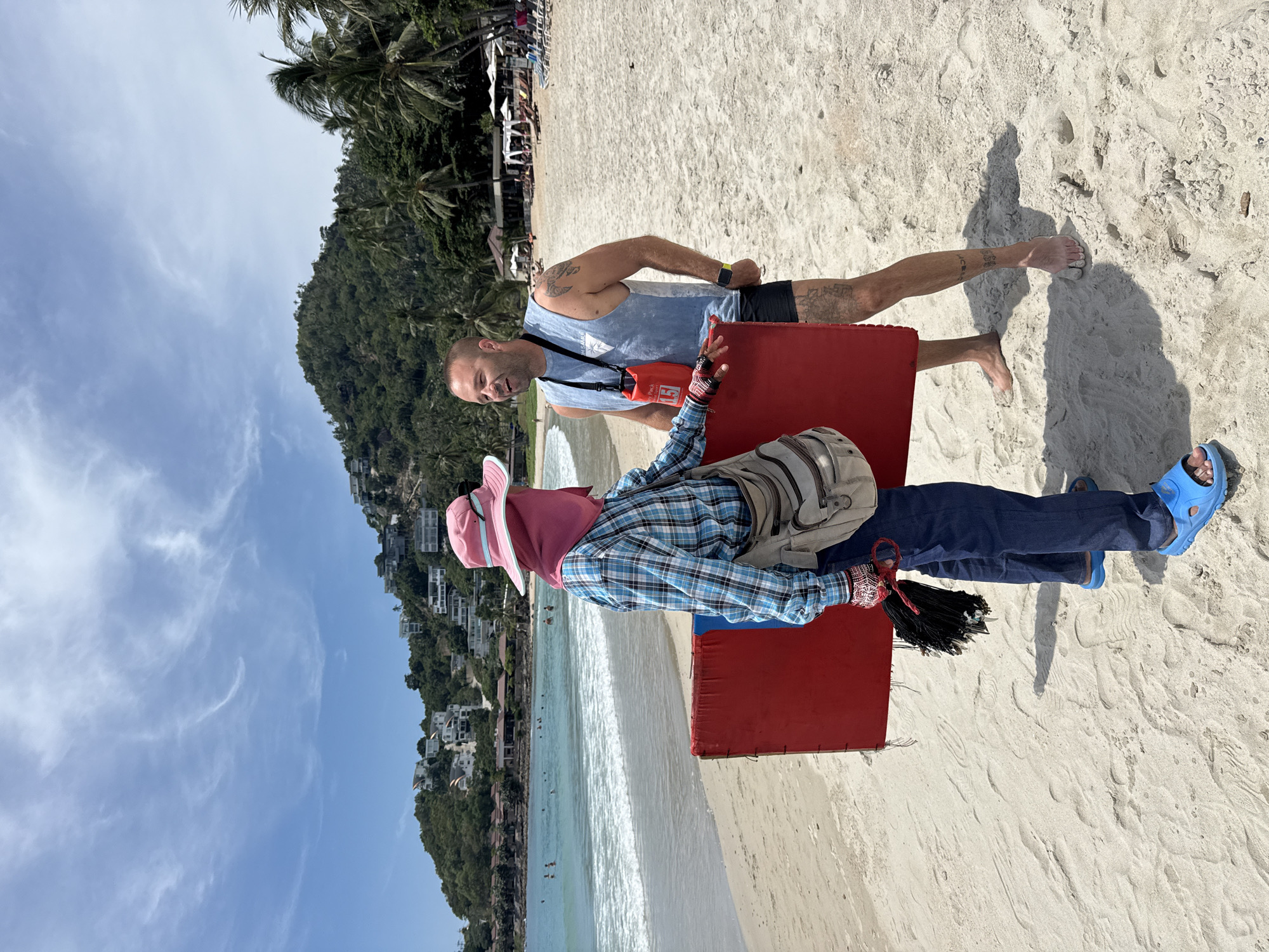 The image captures a beach scene on the coast of Ko Samui District, Thailand. Two individuals are the central focus. On the left, a person wearing a pink wide-brimmed hat with a face covering, a blue and white checkered long-sleeved shirt, and blue jeans stands partially turned to the right. They are holding a large, flat red object with a blue edge. A light-colored messenger bag is slung over their shoulder, and they are wearing blue sandals. They also appear to be holding a bundle of black objects in their gloved hands.
To the right, a man with short hair and tattoos on his arms and legs is facing the person with the red object and smiling. He is wearing a light blue sleeveless shirt with text on it and dark shorts. A red waterproof bag with a strap is hanging from his shoulder. He is barefoot, with one foot slightly in the sand.
The setting is a sandy beach with the ocean visible in the background. Gentle waves are breaking on the shore. Beyond the beach, lush green hills rise, dotted with white buildings, suggesting a developed resort area. Palm trees are scattered throughout the landscape, and some lounge chairs and umbrellas are visible further down the beach. The sky is partly cloudy with scattered white clouds against a blue expanse. The overall impression is a sunny day on a tropical beach.