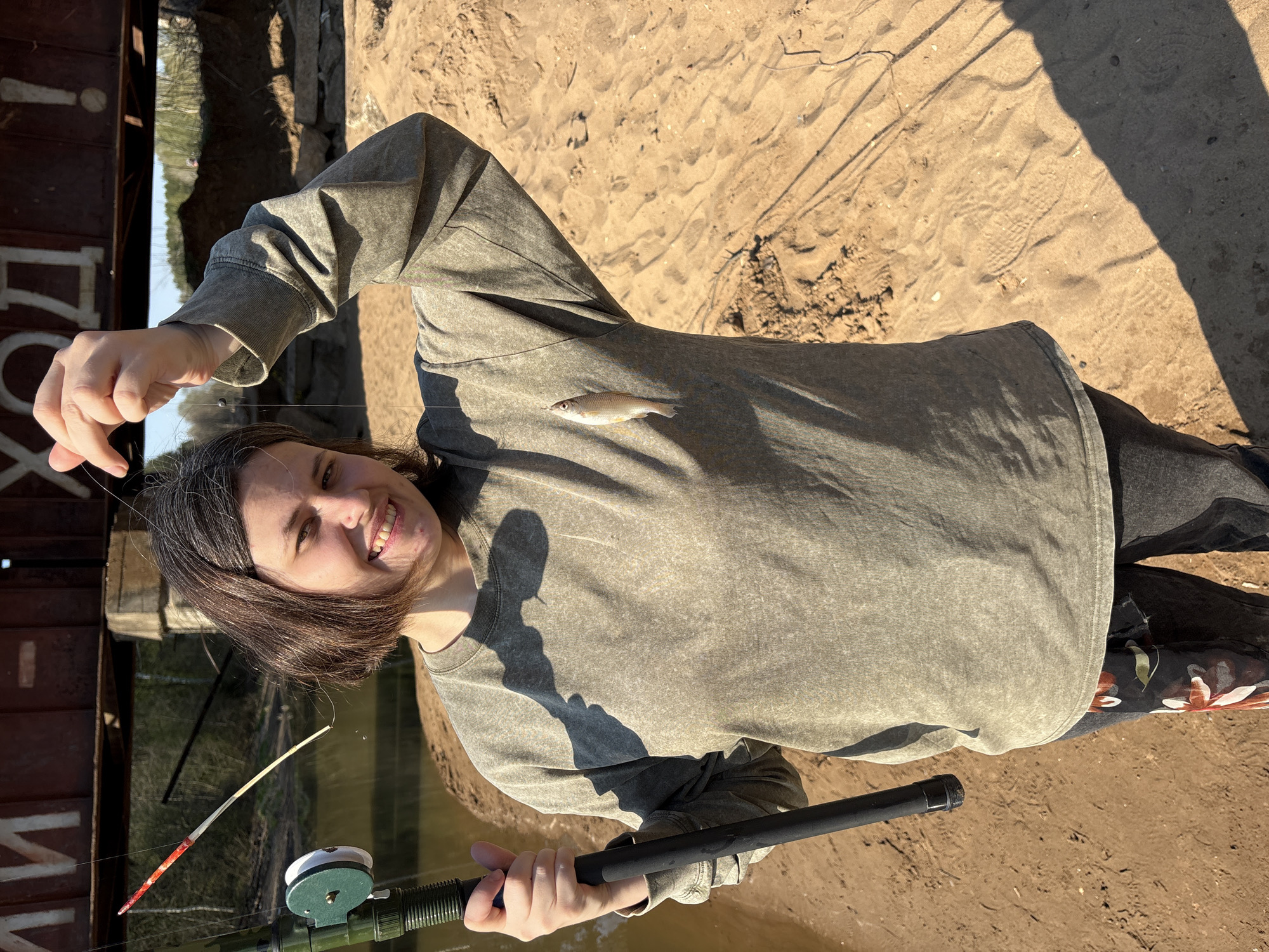 A woman is standing by the Kama River in Chaikovsky, Russia, holding a fishing rod and a small fish. She is smiling at the camera. The background includes the river, sandy banks, and a structure that might be part of a bridge. The woman is wearing a gray sweatshirt and dark pants. Her fishing rod has a green reel and a red and white tip. The fish hangs from the line in front of her shirt. The scene is brightly lit by sunlight.