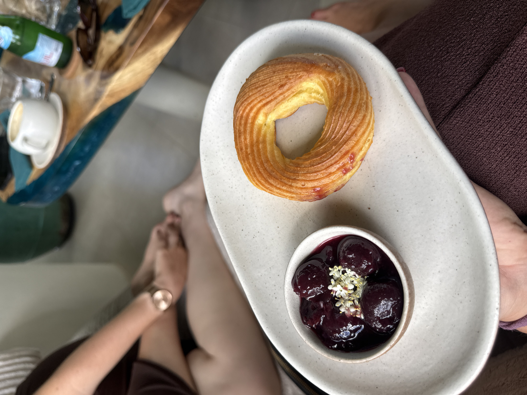 A high-angle medium shot depicts a white elongated plate resting on the lap of a person wearing brown clothing. The plate holds two items: a circular pastry with ridges and a small white bowl filled with dark cherries and white flowers. A woman's arm with a gold watch is visible to the left. A table with a coffee cup, a water bottle, and sunglasses is slightly out of focus in the background. The location is North Kuta, Indonesia.