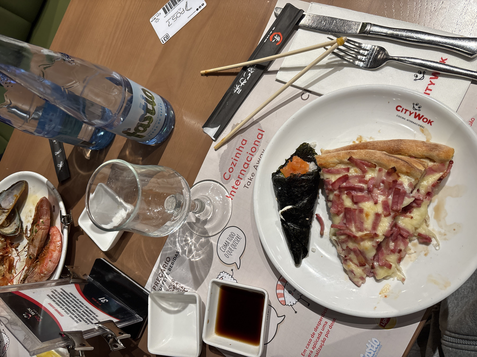 The image depicts a dining table scene, likely in a restaurant located in Vila Nova de Gaia, Portugal, identifiable from the "Fastio" water bottle, a brand of water from Serra do Gerês, Portugal. The table is cluttered with various items, including a bottle of water, an empty wine glass, a plate with a piece of pizza and a Temaki sushi, a plate of seafood (shrimp and mussels), chopsticks in a paper wrapper, cutlery (knife and fork), small square dishes (one empty and one with soy sauce), menus, and a table card with the number 31. The restaurant branding "City Wok - Cozinha Internacional" is visible on the plate and menu, suggesting an Asian-themed cuisine. The presence of a bill/receipt and used dishes indicates that a meal is in progress or has recently concluded. The environment is indoors, with the table surface appearing to be made of wood.