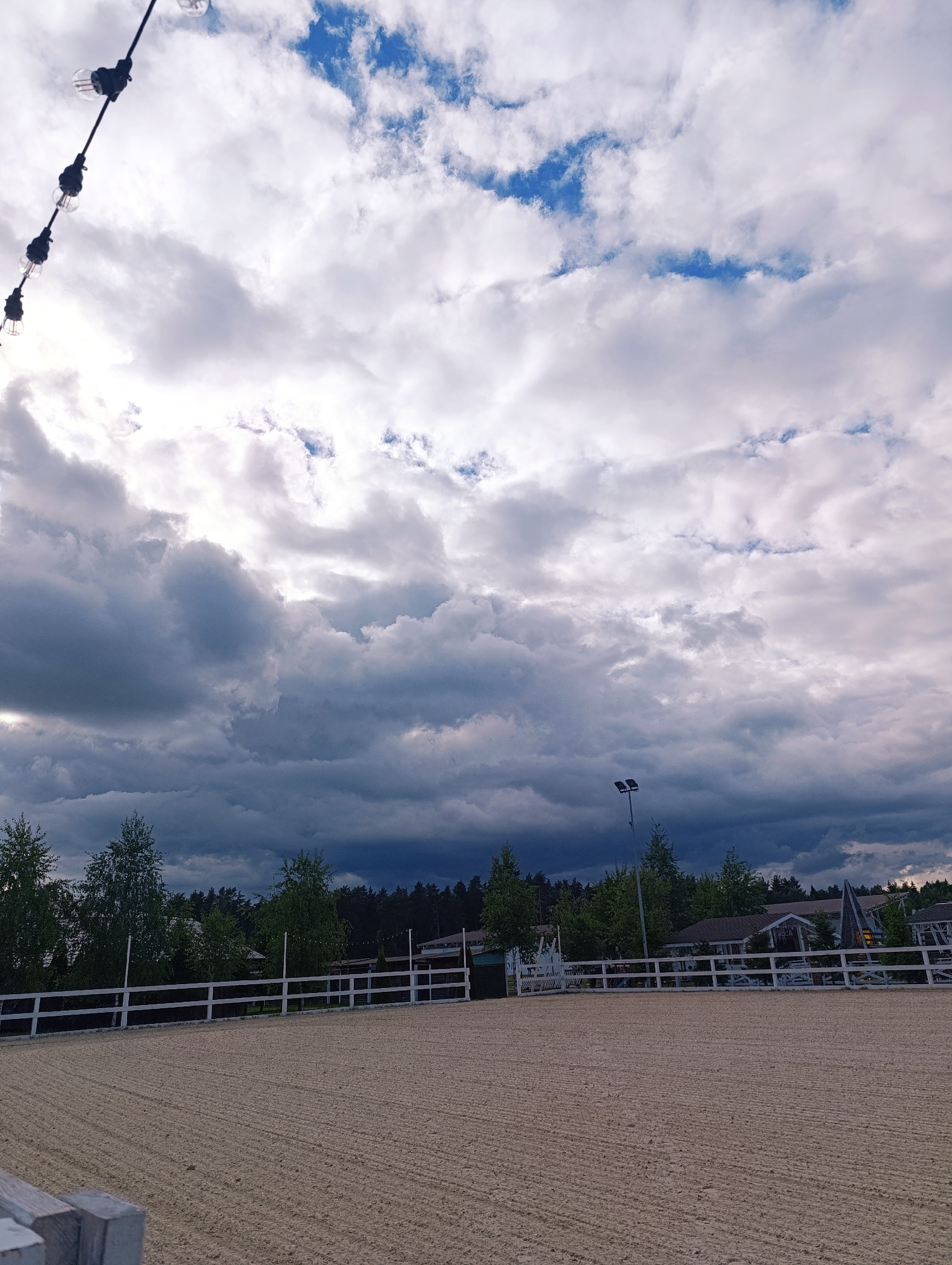 The image shows an outdoor riding arena in Ivanovskoe, Russia. The arena is enclosed by a white wooden fence, with a sand surface marked by lines from dragging. Trees and buildings are visible behind the fence. The sky is filled with dark storm clouds, with patches of blue visible between them. A string of lights hangs in the upper left corner. A light pole is visible in the middle distance.