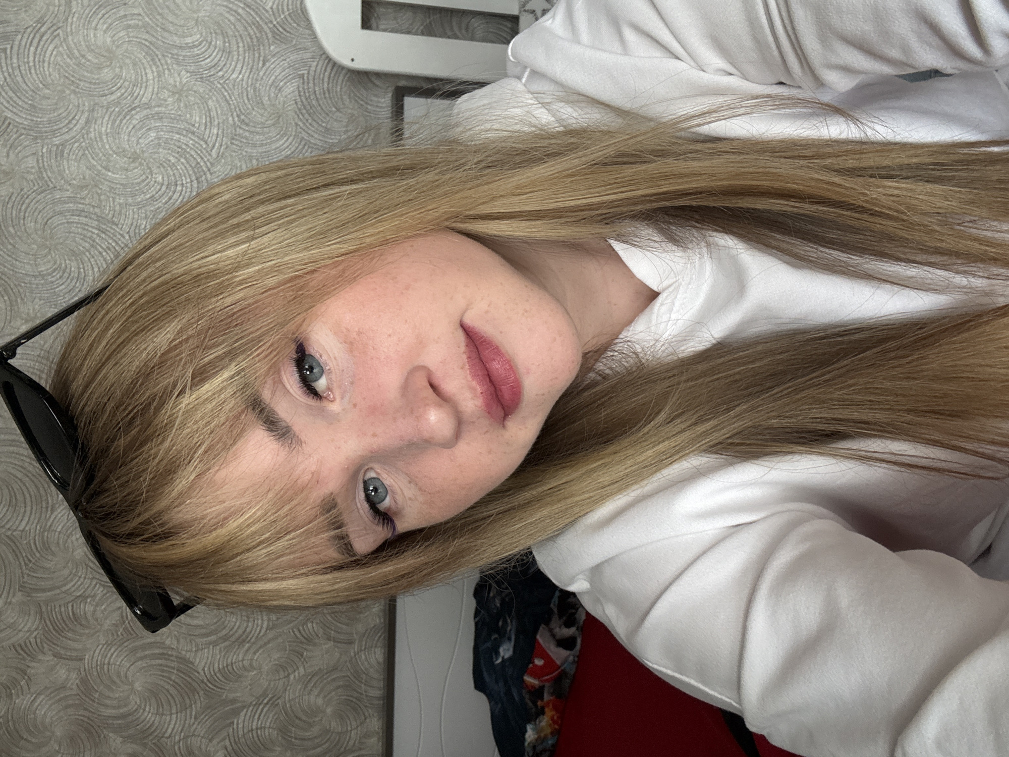 A young woman with blonde hair, light skin, and blue eyes is looking at the camera. She has visible freckles on her nose and cheeks. Her hair is styled with bangs. She is wearing a white hooded sweatshirt. Sunglasses are placed on her hair, near the left side of the frame. The background is a textured gray wallpaper. A white object is visible in the upper part of the image. The scene appears to be indoors, possibly a room with a white door in the lower part of the frame. The location is Марковский, Russia.