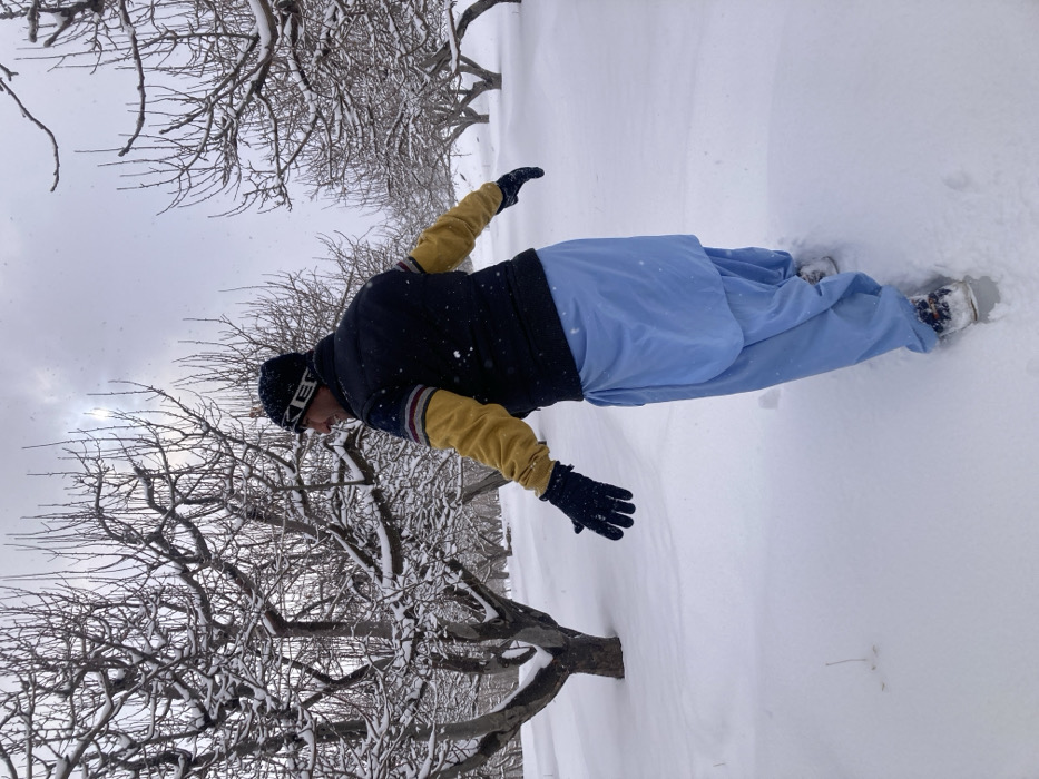 A male individual is depicted walking through a snow-covered orchard in Kanozai, Pakistan, on an overcast day with light snowfall. The individual, seen from behind and slightly to the right, is looking back over his right shoulder with a smile. He wears a black beanie, a dark vest over a long-sleeved shirt with mustard yellow sleeves and striped shoulders, dark blue gloves, and light blue shalwar, with snow-covered footwear. His arms are slightly outstretched to the sides as he traverses the deep snow, leaving footprints. The environment consists of numerous bare deciduous trees, heavily laden with snow, creating a patterned landscape under a cloudy sky with a diffused sun visible in the upper left quadrant.