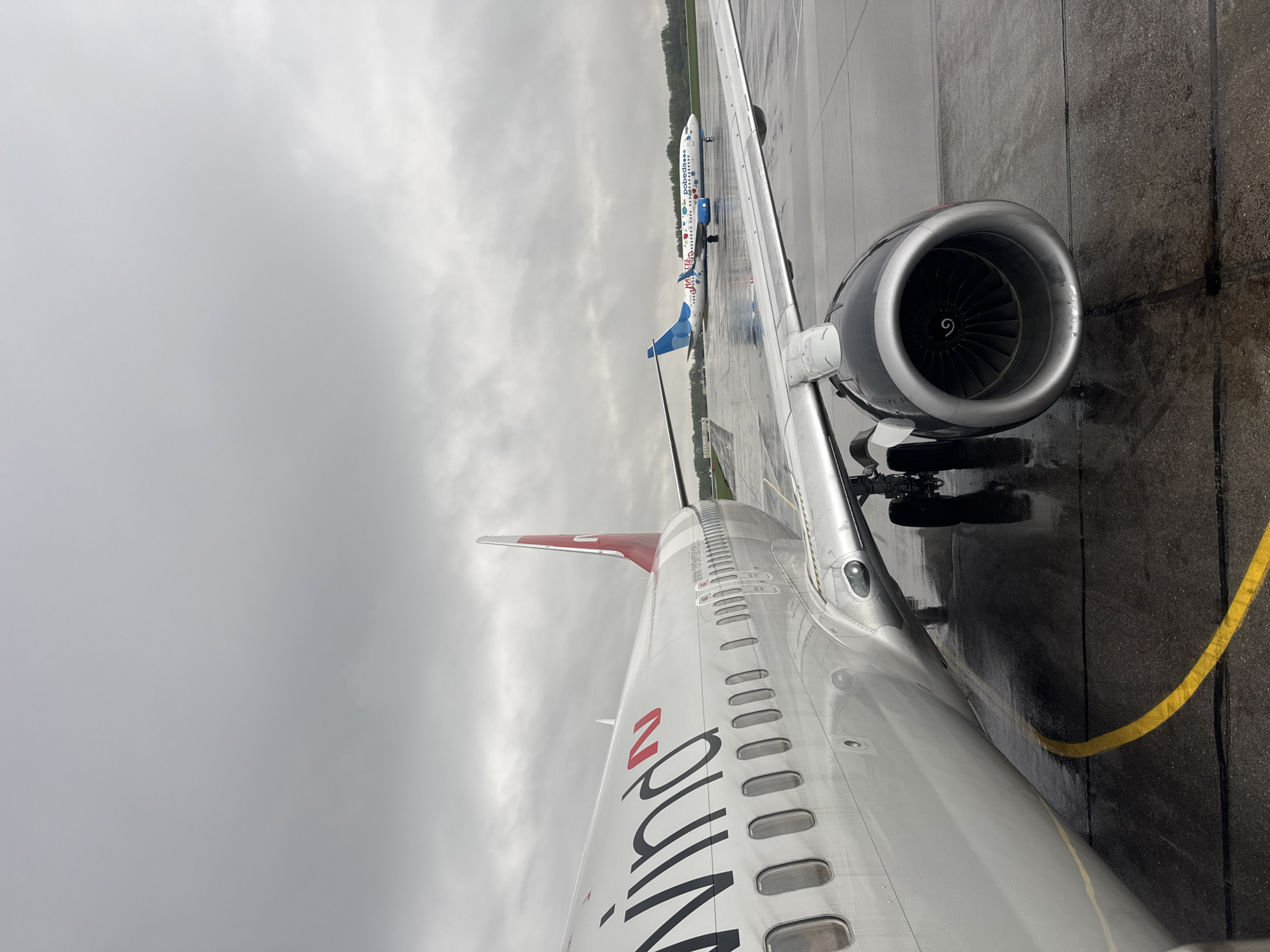 The image shows a view from inside an airplane at Bolshoye Savino Airport in Perm, Russia. The wing and engine of the airplane are visible, along with another plane in the distance. The ground is wet, suggesting it has been raining. The sky is overcast. The name "wing" is visible on the side of the airplane.