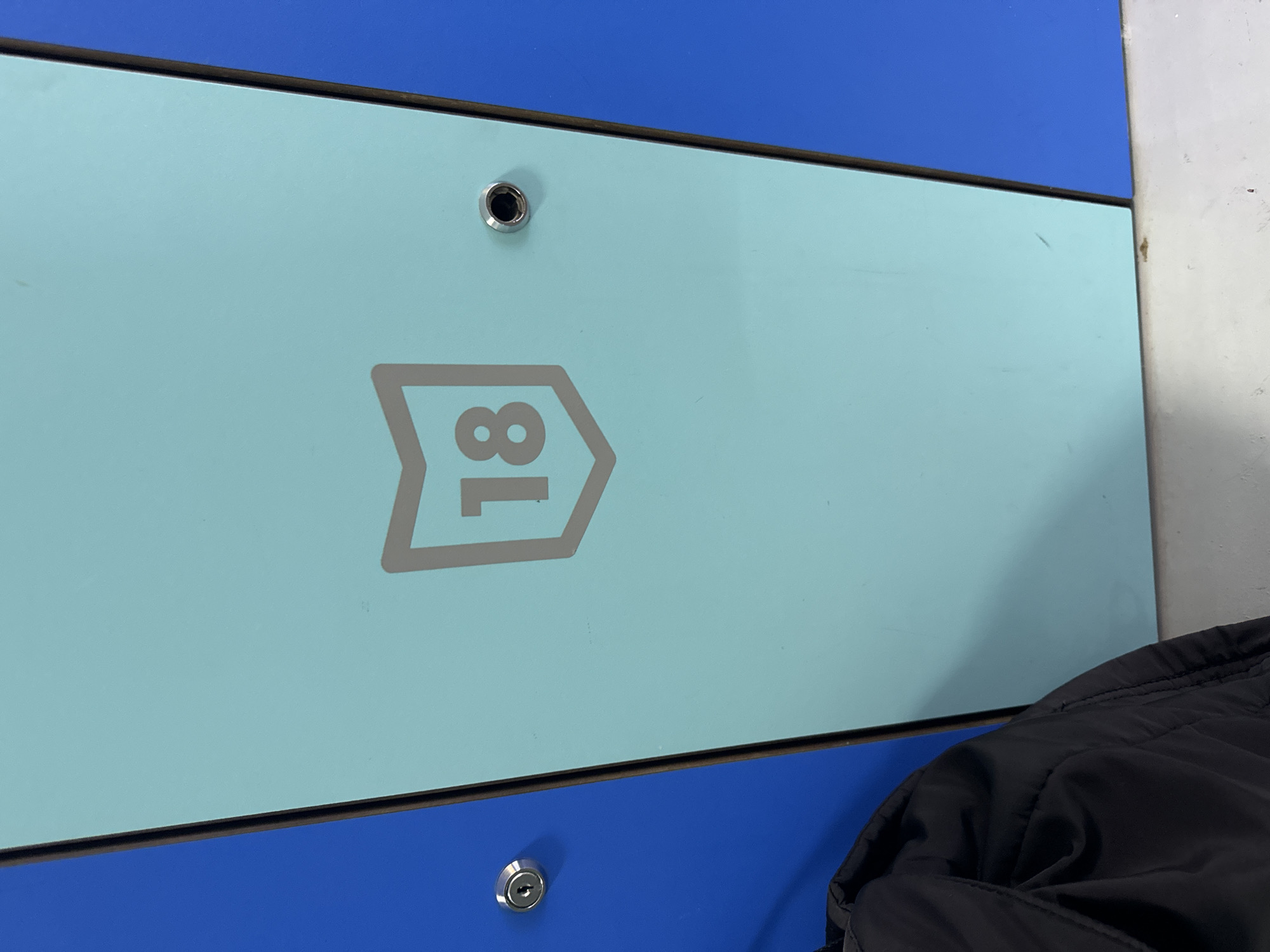 A close-up, eye-level shot shows a light blue locker door with a gray shield symbol containing the number "18" in gray. The locker door is flanked by a bright blue locker door on the left and a bright blue surface on the right. A silver lock is visible on the blue locker door to the left. A small silver knob or keyhole is present on the light blue locker door. A black jacket is partially visible in the lower left corner. The scene is likely in a public facility such as a gym or pool changing room. There is no indication of the specific location within the city of Murcia, Spain.