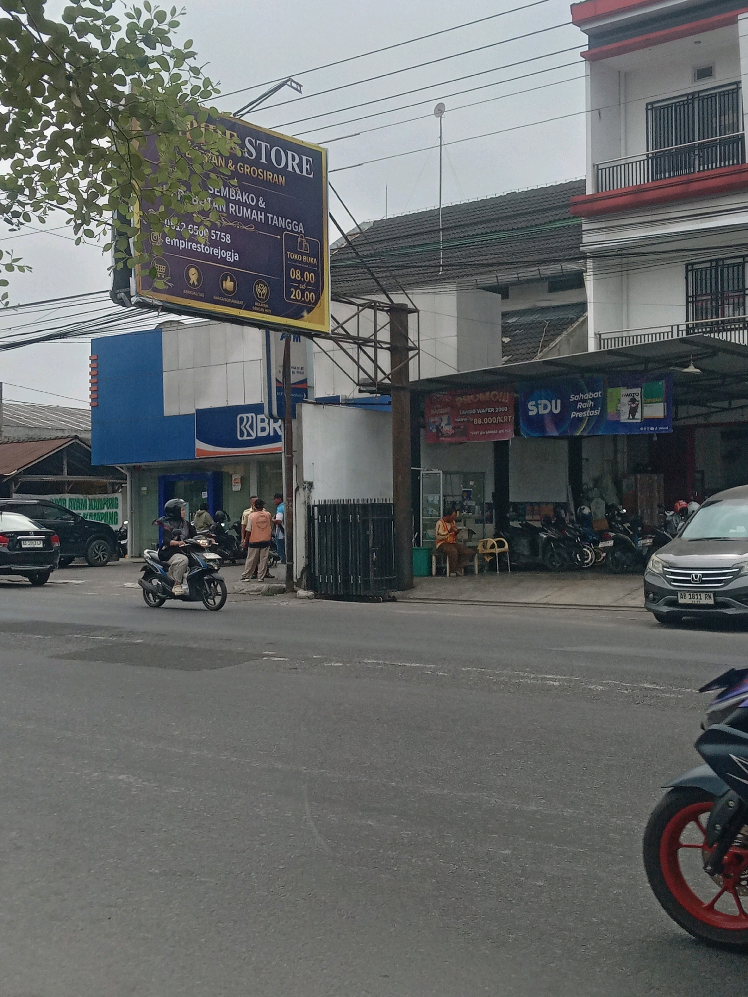 A street scene in Maguwoharjo, Indonesia, features a billboard for "EMPIRE STORE" hanging from a metal frame, partially obscured by tree branches. The billboard advertises "SEMBAKO & PERALATAN RUMAH TANGGA," or groceries and household appliances, with contact information and operating hours of 08.00 to 20.00. In front of the billboard, a blue building with white accents displays the "BRI" logo, indicating a bank. Several people are visible near the BRI building, some standing and others seated. A motorcycle rider wearing a helmet is on the road, and other motorcycles are parked nearby. The street is paved, and power lines crisscross overhead. To the right, a white building with red trim and balconies stands. A dark gray car with a license plate reading "AB 1811 RN" is parked on the side of the road. Further down the street, another vehicle, a dark SUV, is visible. The sky is overcast.