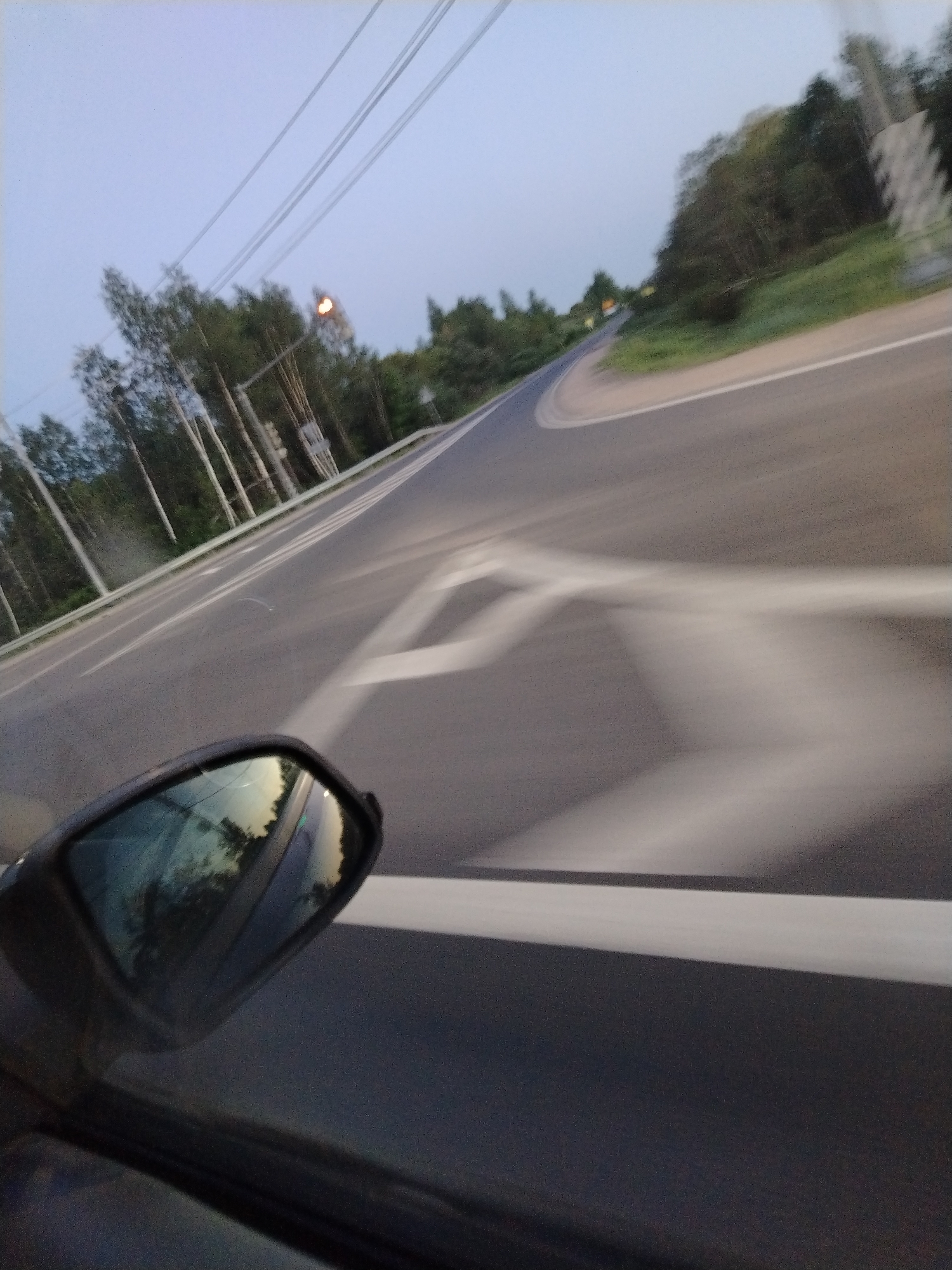 A blurred, upward-angle, low-light photograph taken from inside a moving car traveling on an asphalt road. The road has white lane markings and is surrounded by trees. A car side mirror is in the lower left foreground, reflecting trees and sky. Overhead power lines are visible in the upper left of the frame. Based on the location provided, the photo was taken in or around the city of Семибратово, Russia. No people are visible.