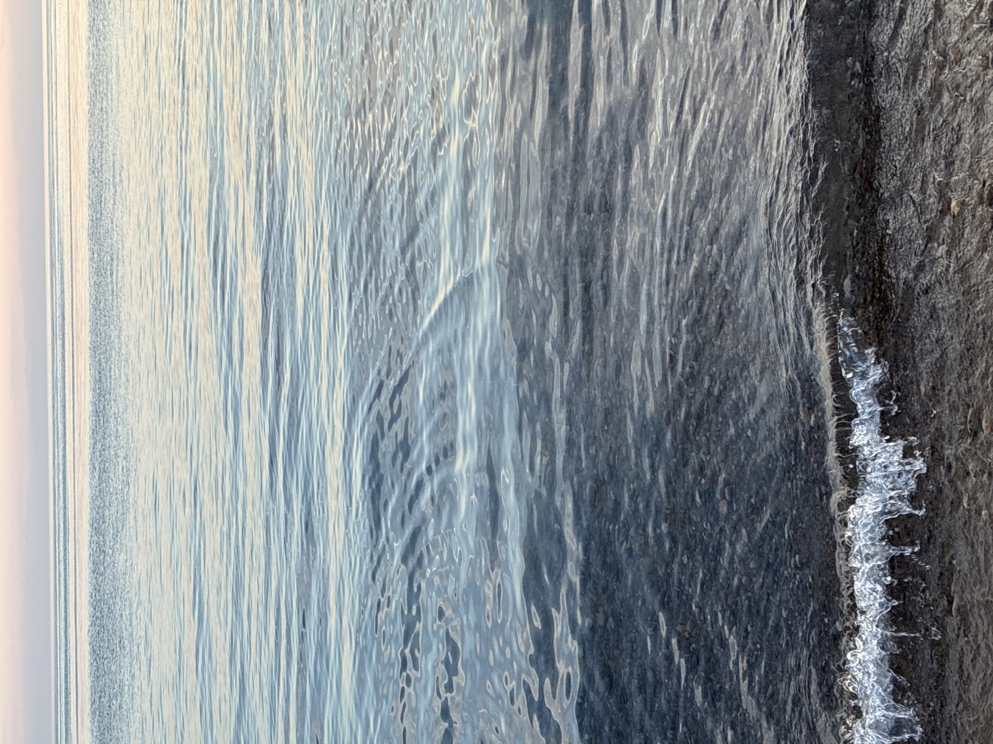 A calm, shallow body of water, likely Lake Baikal in Listvyanka, Russia, is depicted. The water is clear, revealing a dark, rocky bottom. Gentle waves lap at the shore, creating a thin line of white foam. The sky above is a pale pink and blue, suggesting either sunrise or sunset. The overall scene is serene and natural, with no people or man-made objects visible.