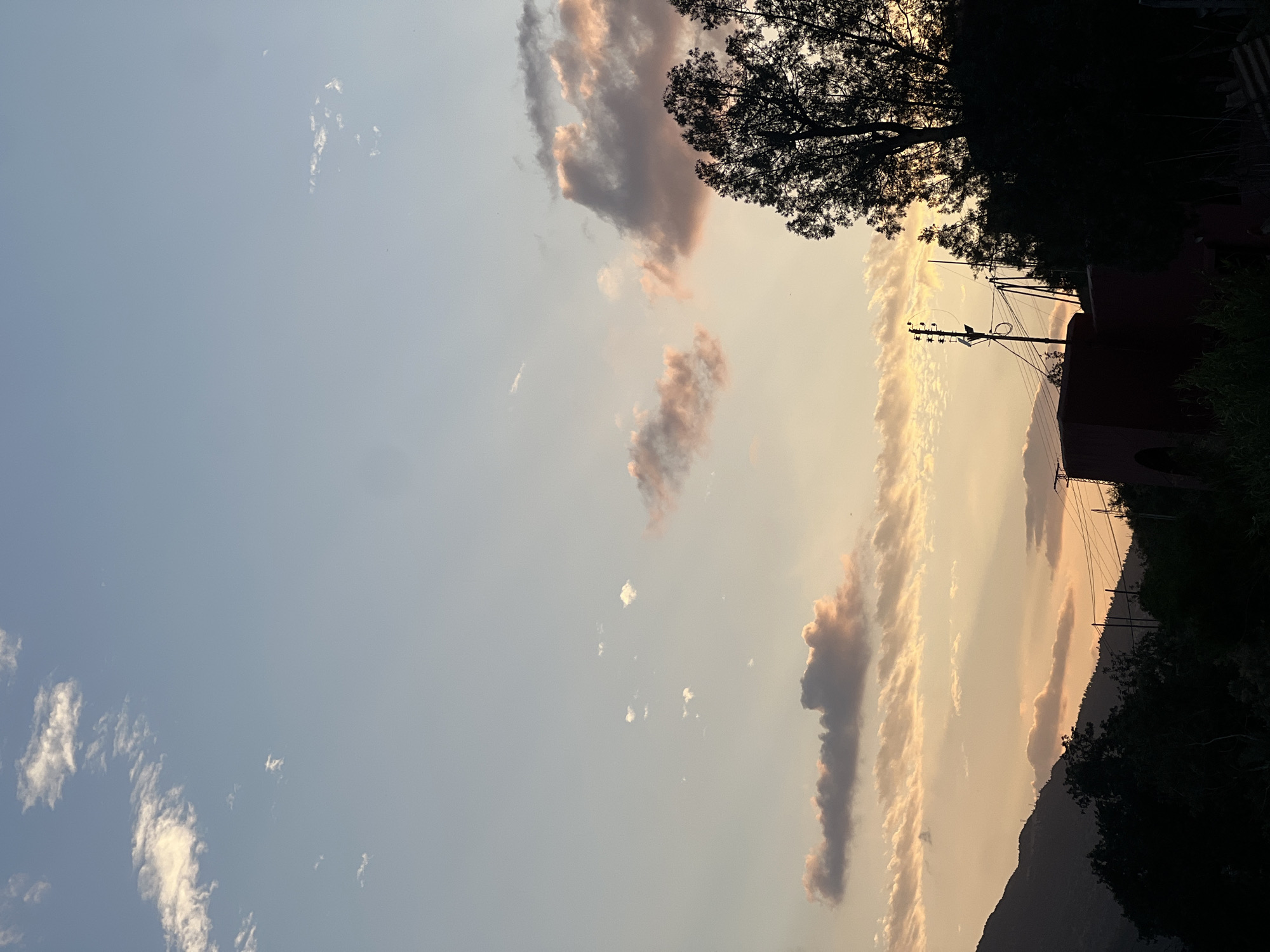 The image depicts a sky scene at sunset in Bin El Ouidane, Morocco. The sky is a mix of pale blue and orange hues, with elongated clouds reflecting the setting sun. Some clouds have a light pinkish tint. Silhouetted trees and a building partially obstruct the view on the right side of the frame. Electrical poles and wires are visible against the colorful sky. The lower part of the frame shows a dark, mountainous landscape.