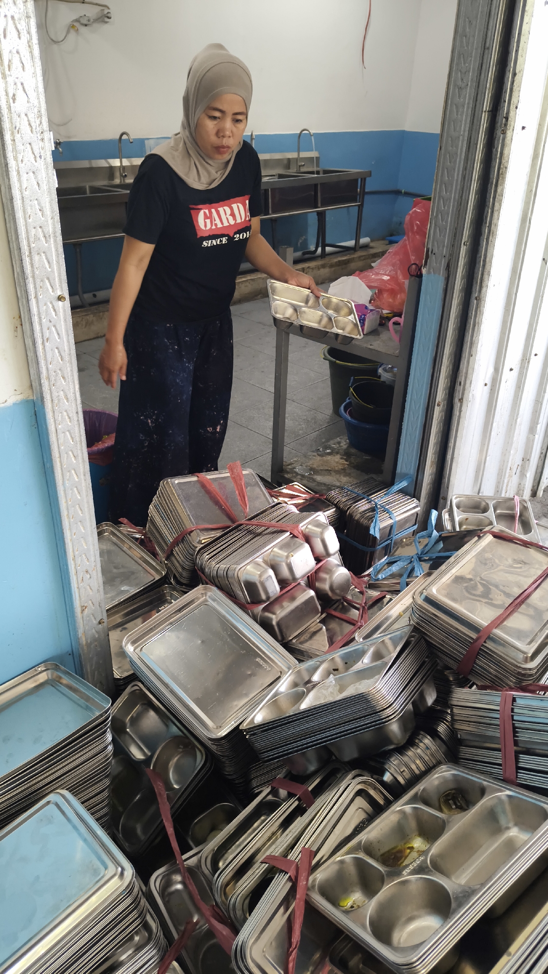 A woman wearing a light brown hijab and a black t-shirt with the word "GARDA" printed on it, stands in a doorway, holding a metal tray with compartments. She is surrounded by large stacks of metal trays, some of which are bundled with red and blue ties. In the background, there are stainless steel sinks and a blue and white tiled wall, suggesting a kitchen or food preparation area. The floor is tiled and appears somewhat wet in places. The overall scene suggests a storage or staging area for food service equipment in what is likely a community setting in Sambipondok, Indonesia.