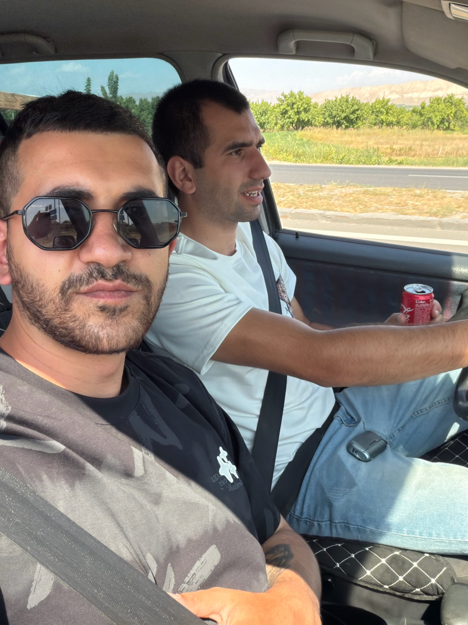 Two men are inside a car driving on a sunny day in the vicinity of Taperakan, Armenia. The man in the foreground, wearing a black t-shirt and sunglasses, is looking towards the camera. The man in the background, who appears to be driving, is wearing a white t-shirt and is holding a can of Coca-Cola. The car's interior is visible, along with a landscape of trees and fields through the windows. The road appears to be paved, and there are mountains in the distance. The overall scene suggests a casual road trip or journey.