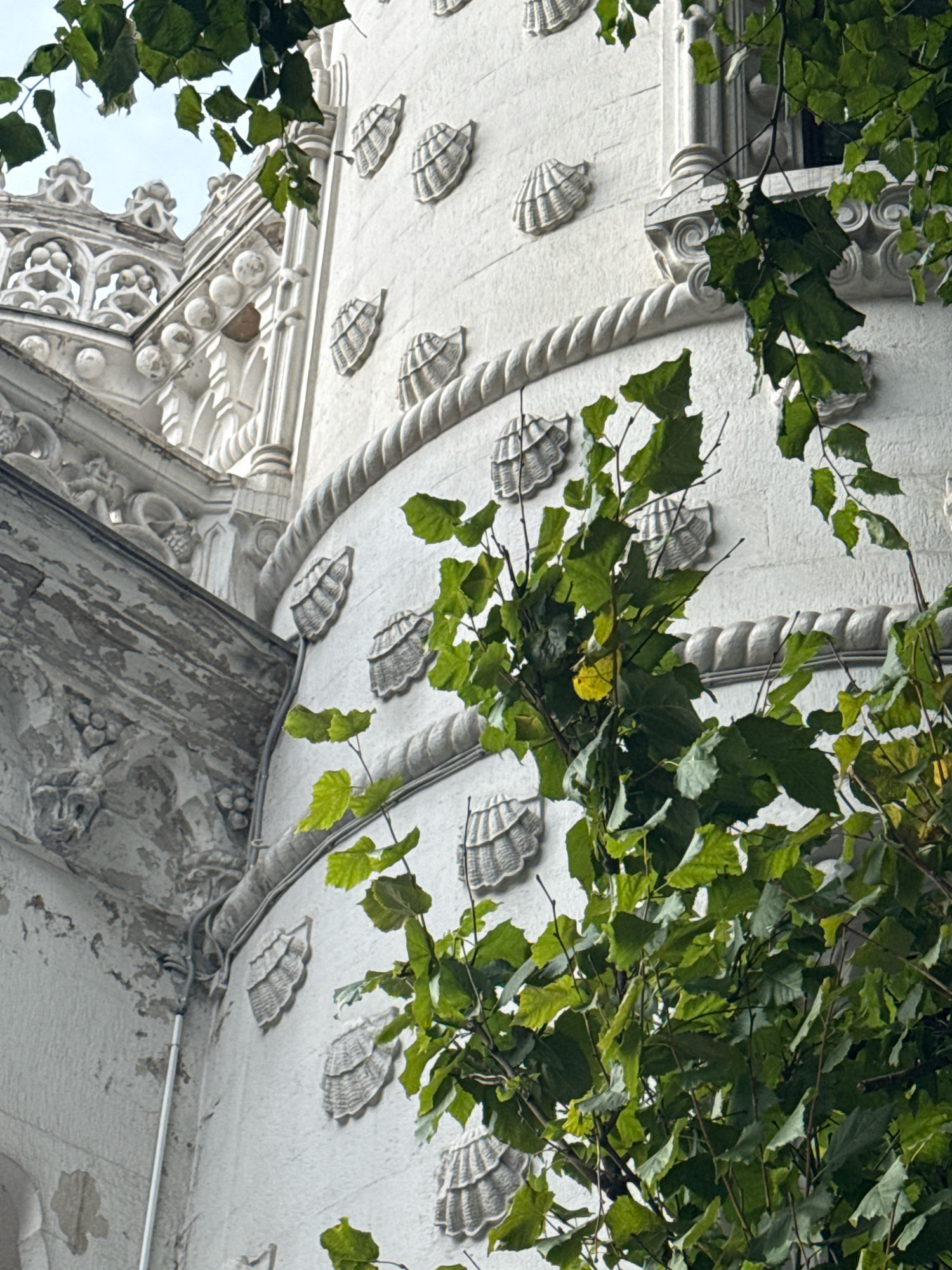 An exterior, eye-level, medium shot shows a white, ornate building facade with shell-shaped embellishments and a rope-like trim, partially obscured by lush green foliage. The building's architecture is intricate, featuring decorative carvings and columns. The sky above is a pale, overcast grey. The scene is static and no people are present.