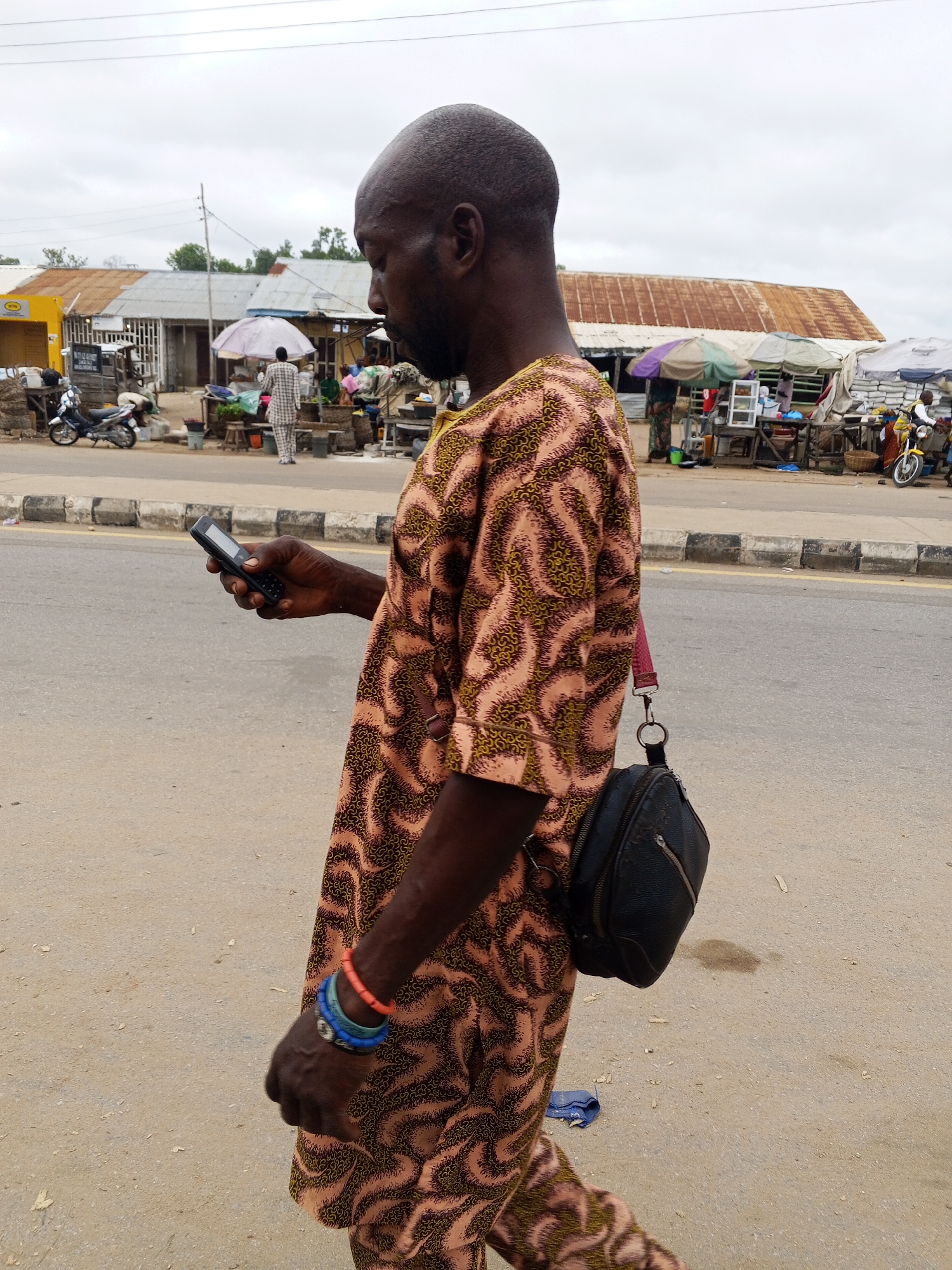 A medium shot shows a bald Black man in a patterned traditional African tunic and blue and orange bracelets on his left wrist, walking on a dirt road in Zango-ndaji, Nigeria. He is holding a black cell phone in his right hand, looking down at it. A black shoulder bag is slung across his right shoulder. In the background, there is a marketplace with stalls covered by umbrellas and corrugated metal roofs. A few motorcycles are parked near the stalls. The sky is overcast.