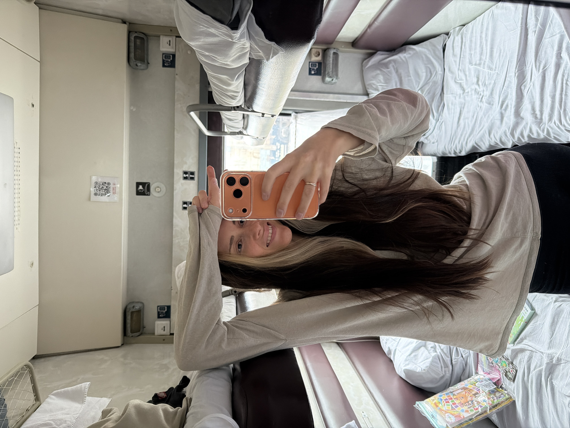 The image is a selfie taken by a young woman inside what appears to be a train sleeper car, specifically a Russian train given the Uzunovo context. The photograph is captured in a mirror or reflective surface, showing the woman's reflection. She is smiling broadly at the camera, her face framed by long, dark brown hair with blonde or caramel highlights. She wears a light gray long-sleeved top and dark leggings. A ring is visible on her left ring finger. She holds an orange or coral-colored smartphone with a visible multi-lens camera setup.
The setting is a compact train compartment. To the woman's right, a neatly made bunk bed with white sheets and a pillow is visible. Above her, a metal frame, likely part of another bunk or a luggage rack, is partially obscured by light-colored fabric. The walls of the cabin are a clean, light cream or white color. Several fixtures are mounted on the wall, including a small light, power outlets, and control panels. A small white sticker featuring a QR code is also visible on the wall, though its text content is unreadable. On a lower surface, possibly a bunk or small table, a colorful packaged item, potentially snacks or a children's activity pack, can be seen. The overall impression is that of a comfortable but functional travel environment during the daytime.